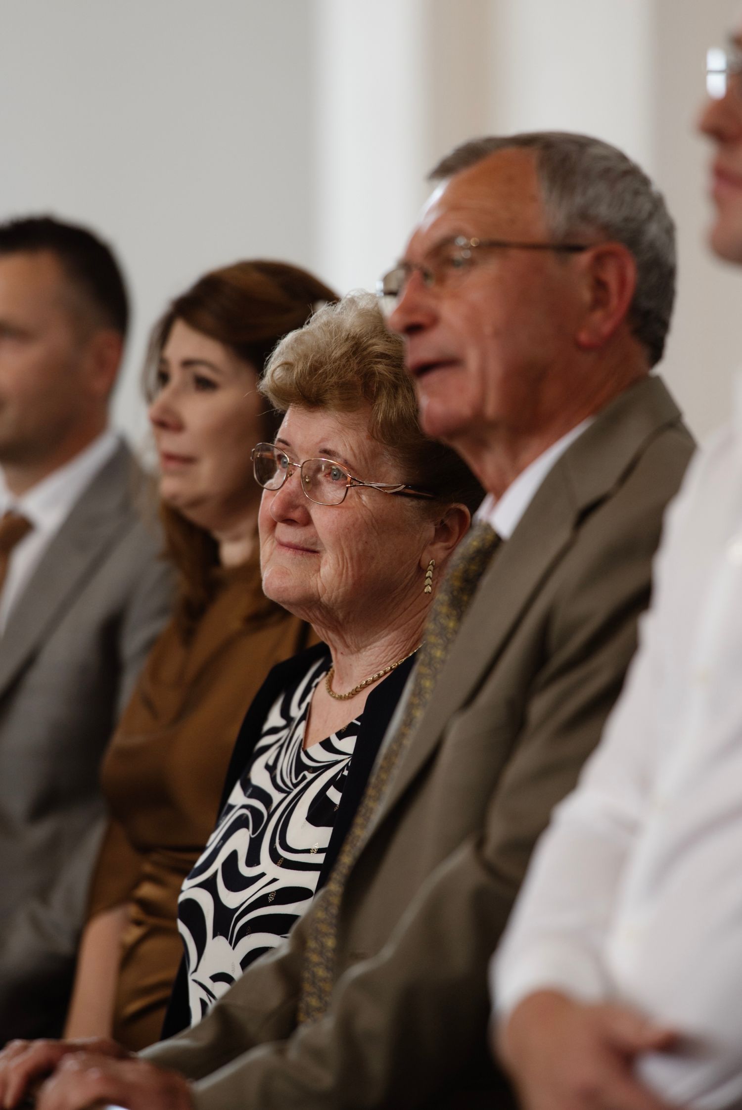 Elderly couple seated at a formal event, woman wearing glasses and floral blouse, man in gray suit behind her.