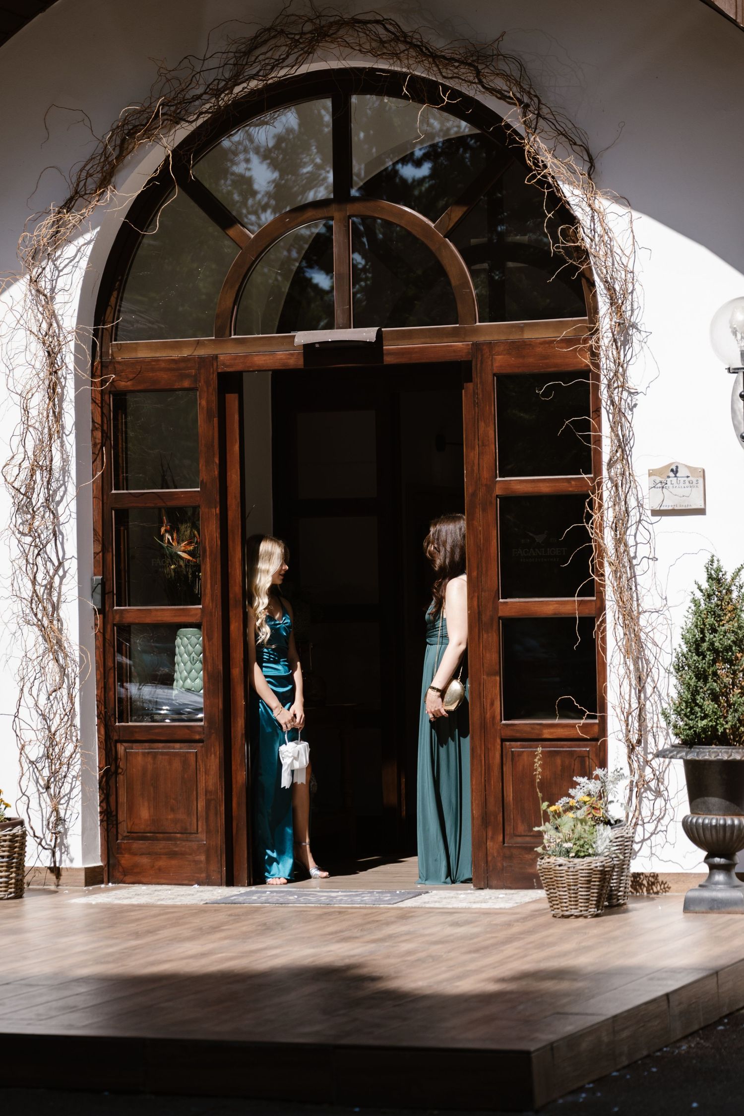 Two women in formal dresses standing in an elegant arched wooden doorway.