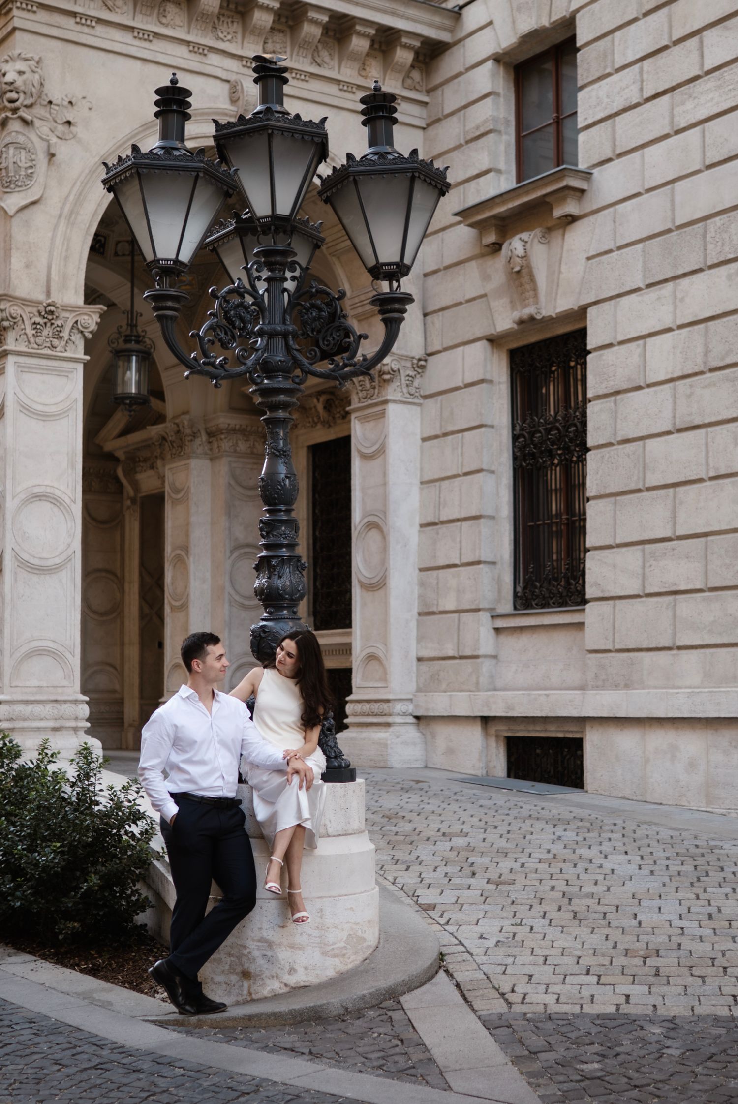 Couple posing by ornate black street lamp outside elegant stone building on cobblestone street.