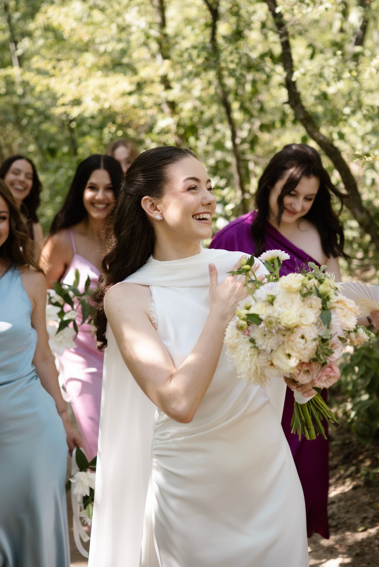 Laughing bride in white gown tossing bouquet with bridesmaids in colorful dresses behind her.