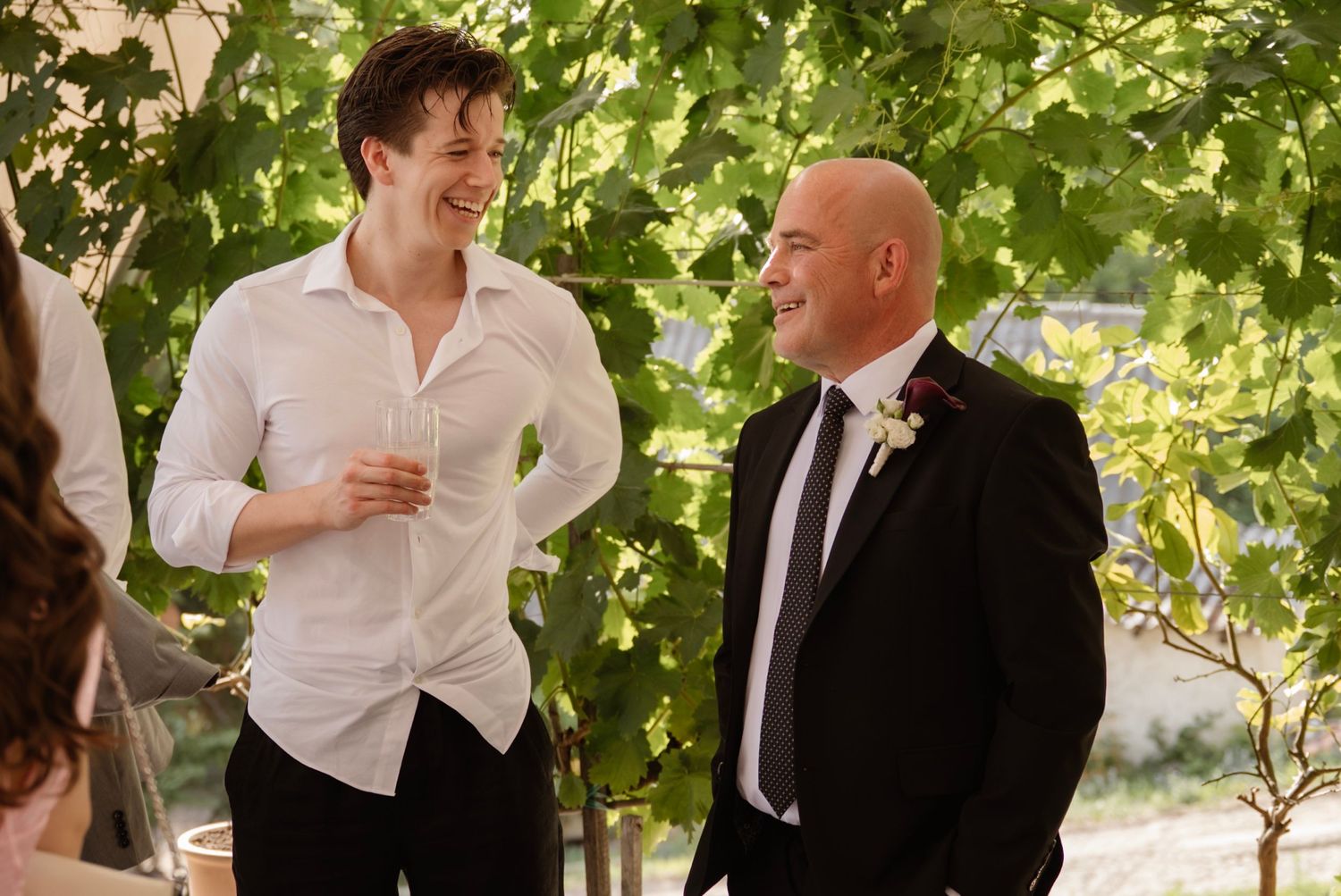 Two men laughing together at an outdoor wedding ceremony surrounded by green vines.