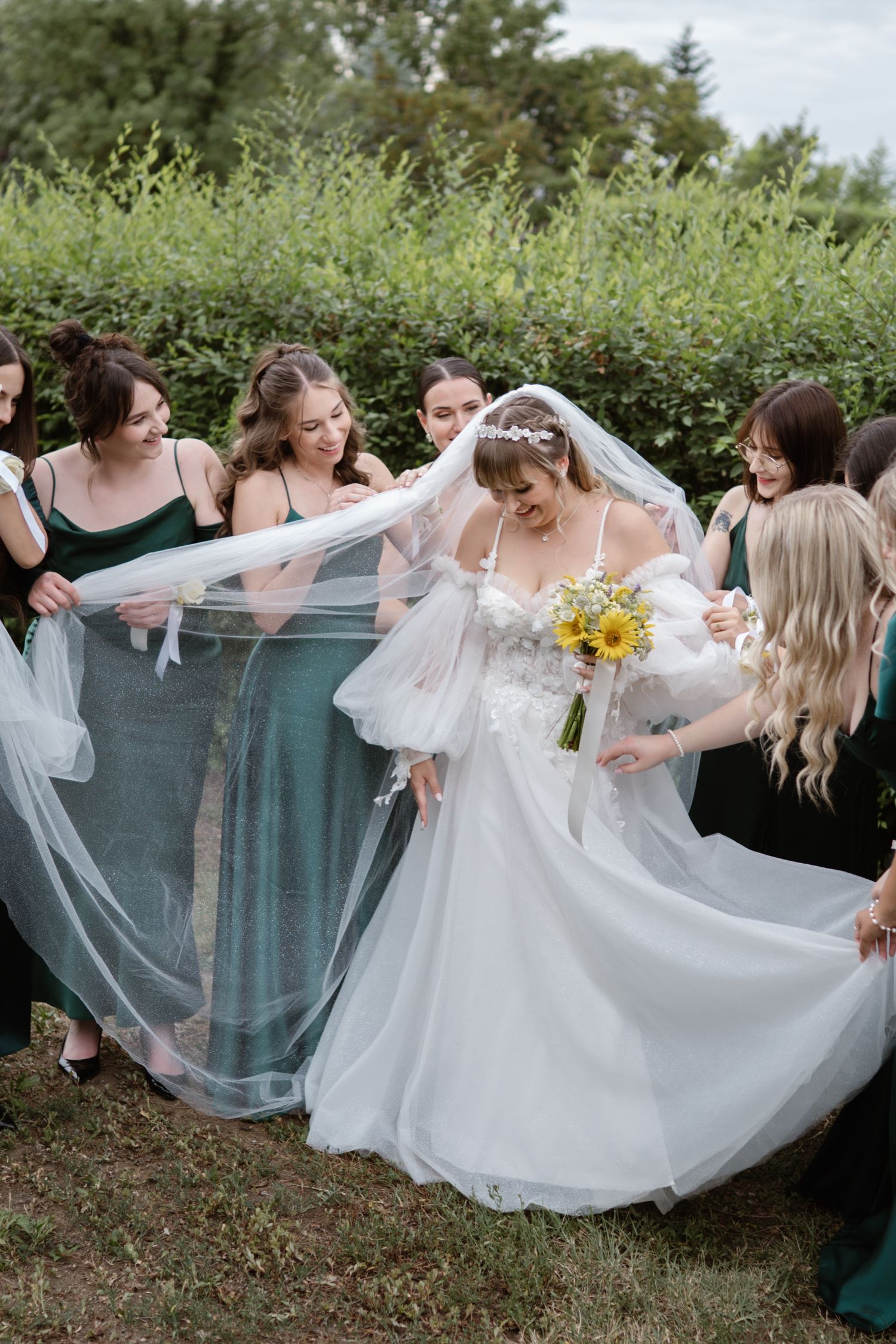 Bridesmaids in green dresses help arrange the bride's flowing veil outdoors.