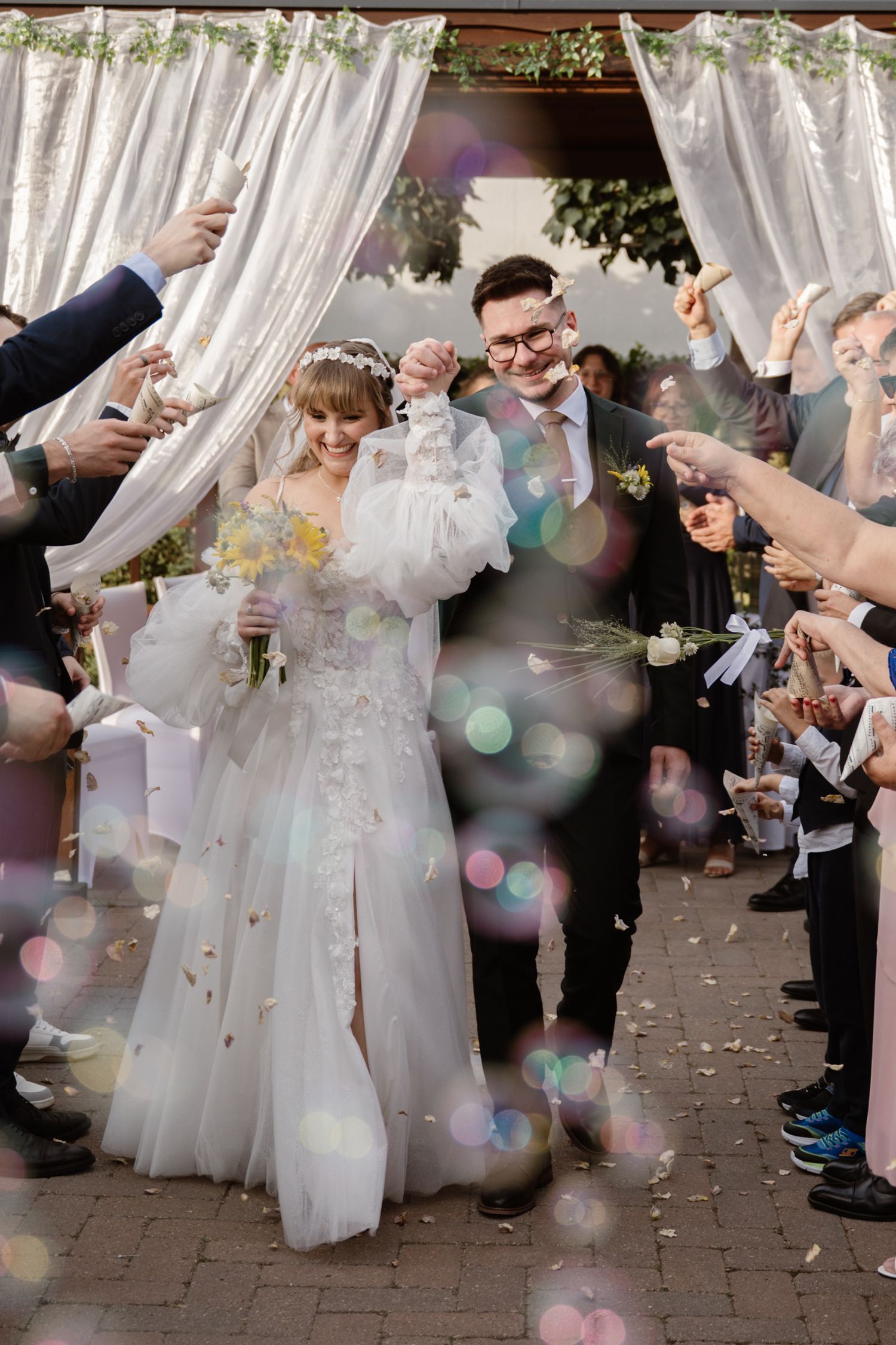 Wedding guests throw confetti as newlyweds walk through a decorated outdoor corridor.