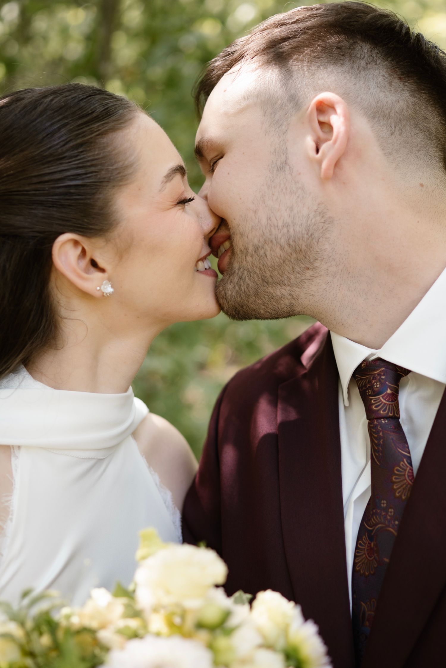A bride and groom share a romantic kiss, dressed in wedding attire with a burgundy suit and white gown, surrounded by greenery.