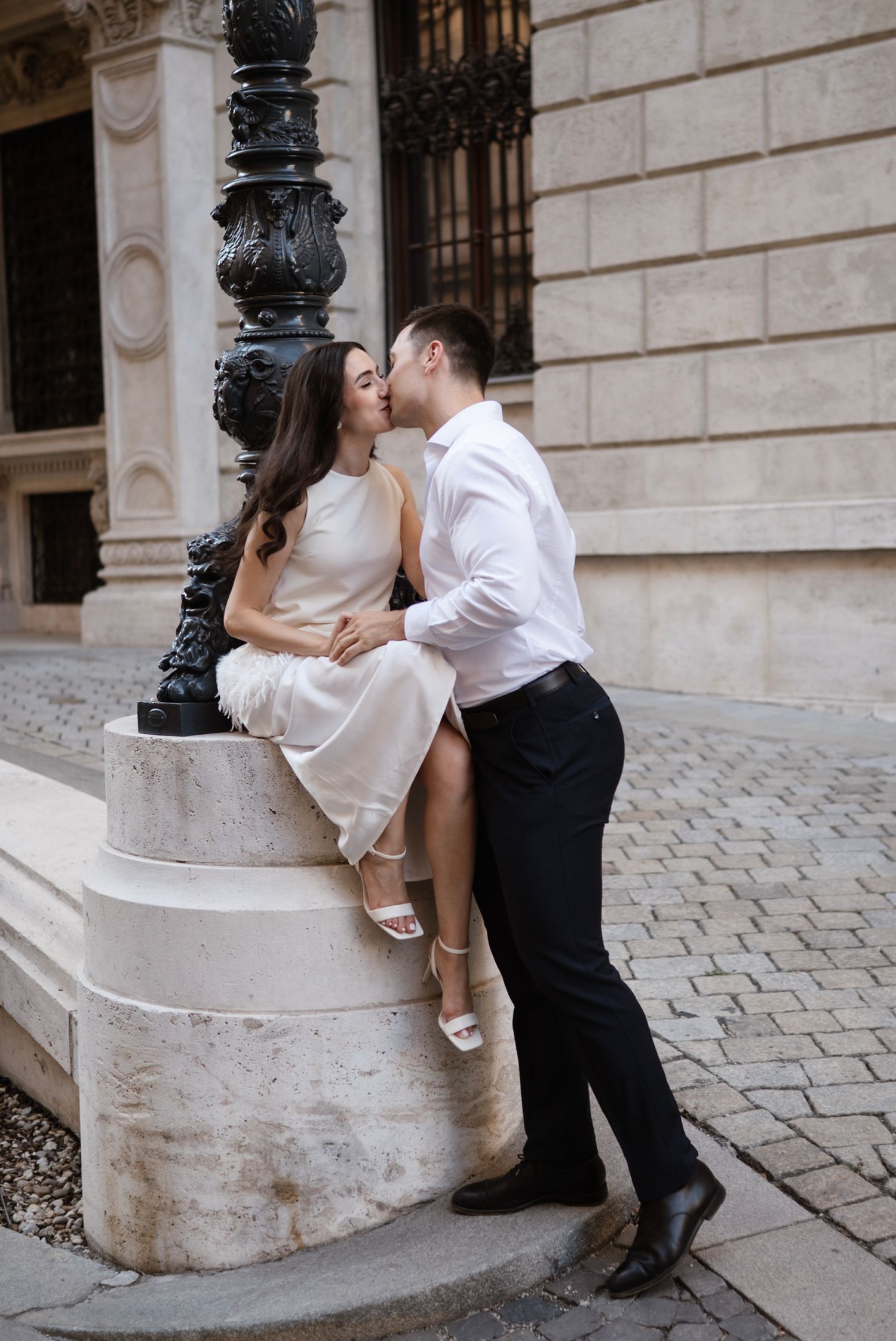 A couple shares a romantic kiss beside an ornate lamppost on a cobblestone European street.