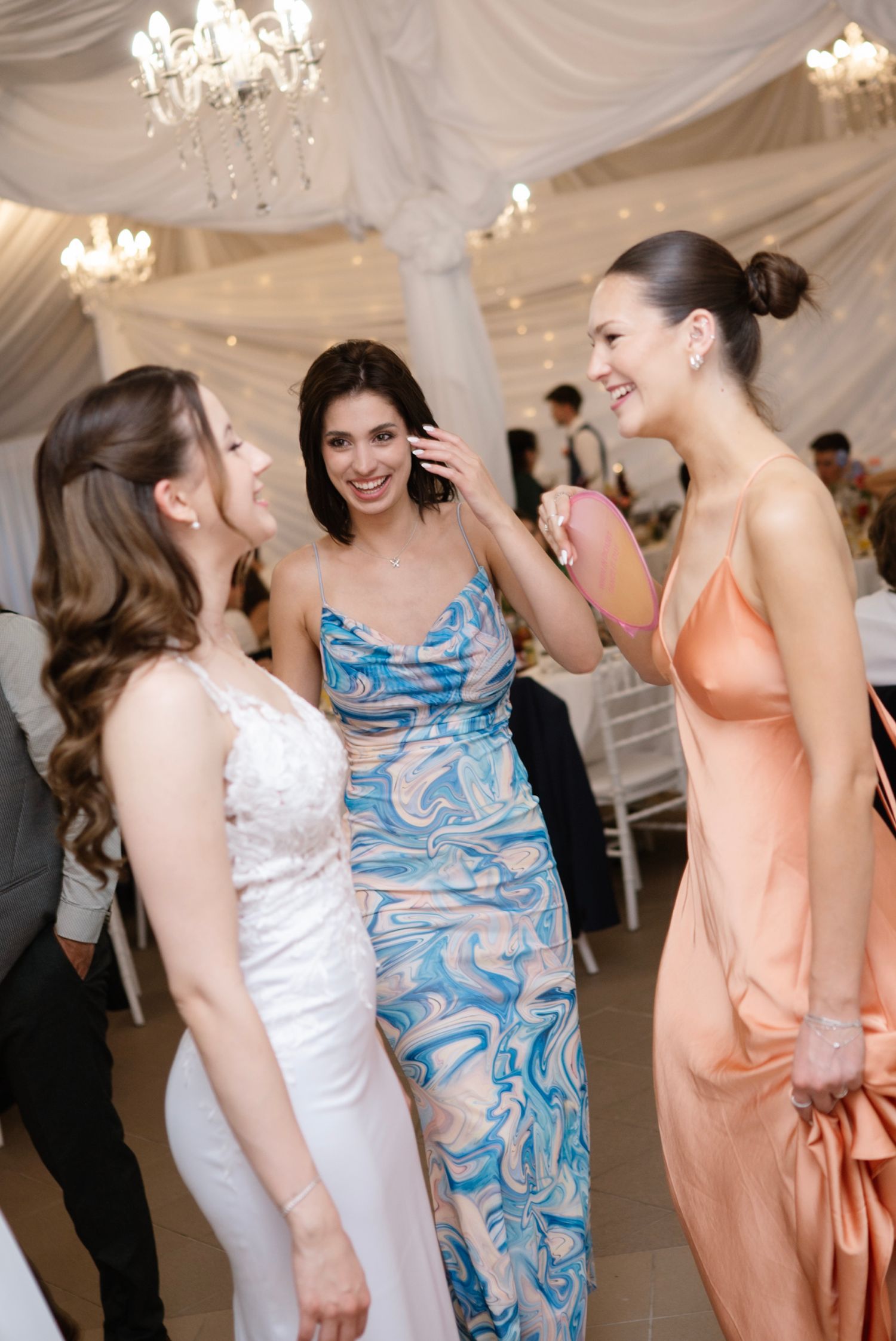 Three elegantly dressed women laugh and chat together at a wedding reception under chandelier lighting.