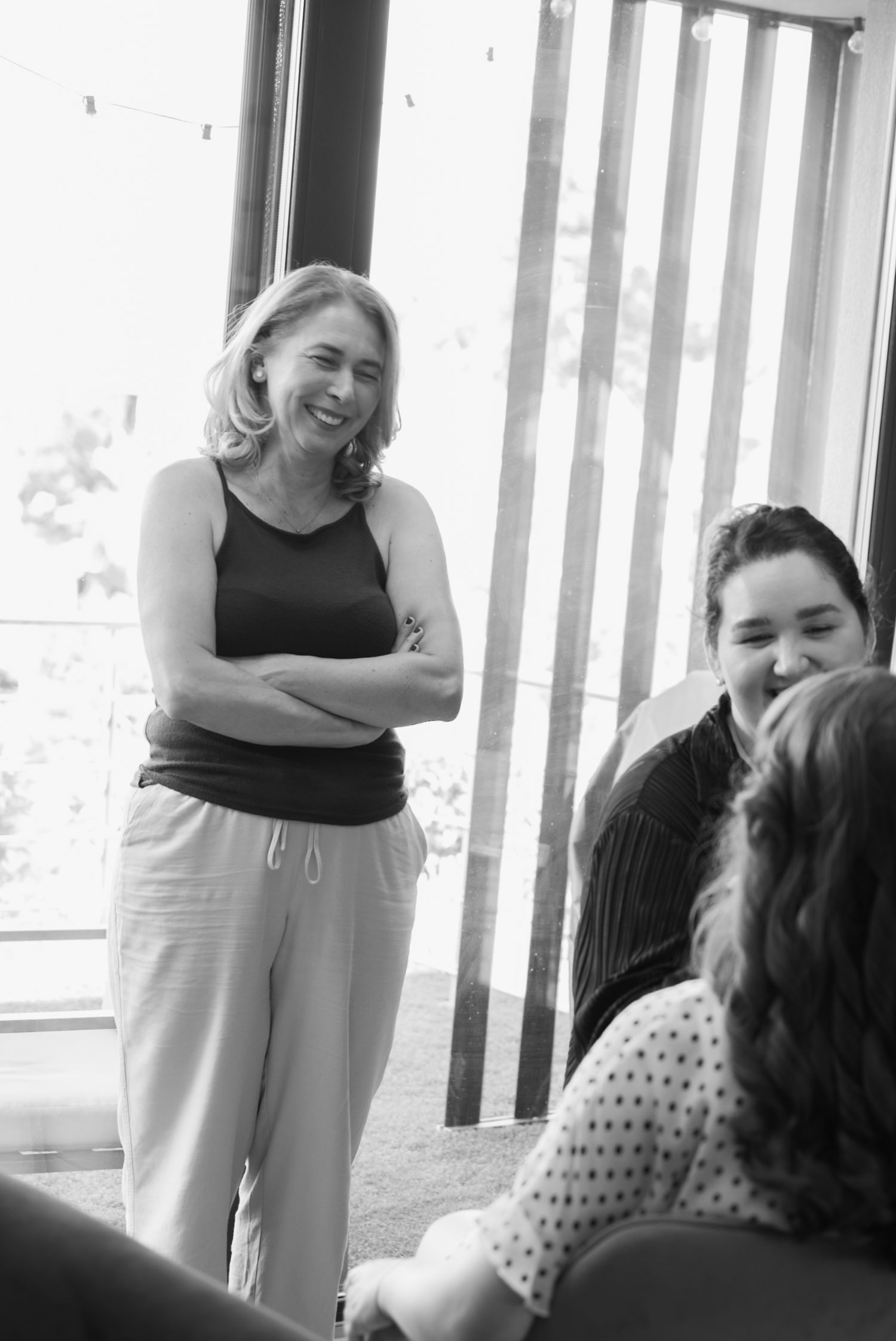 Woman with crossed arms smiling in black and white photo, standing near window with others seated nearby.