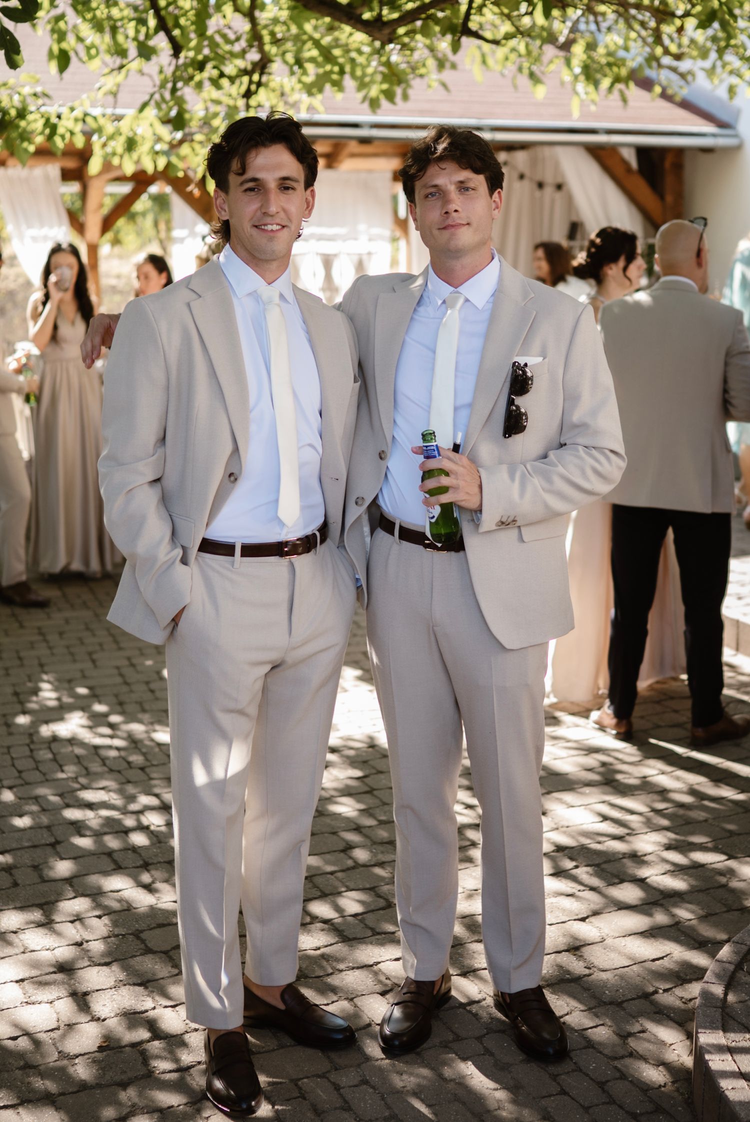 Two groomsmen in matching tan suits and white ties posing outdoors at a wedding reception.
