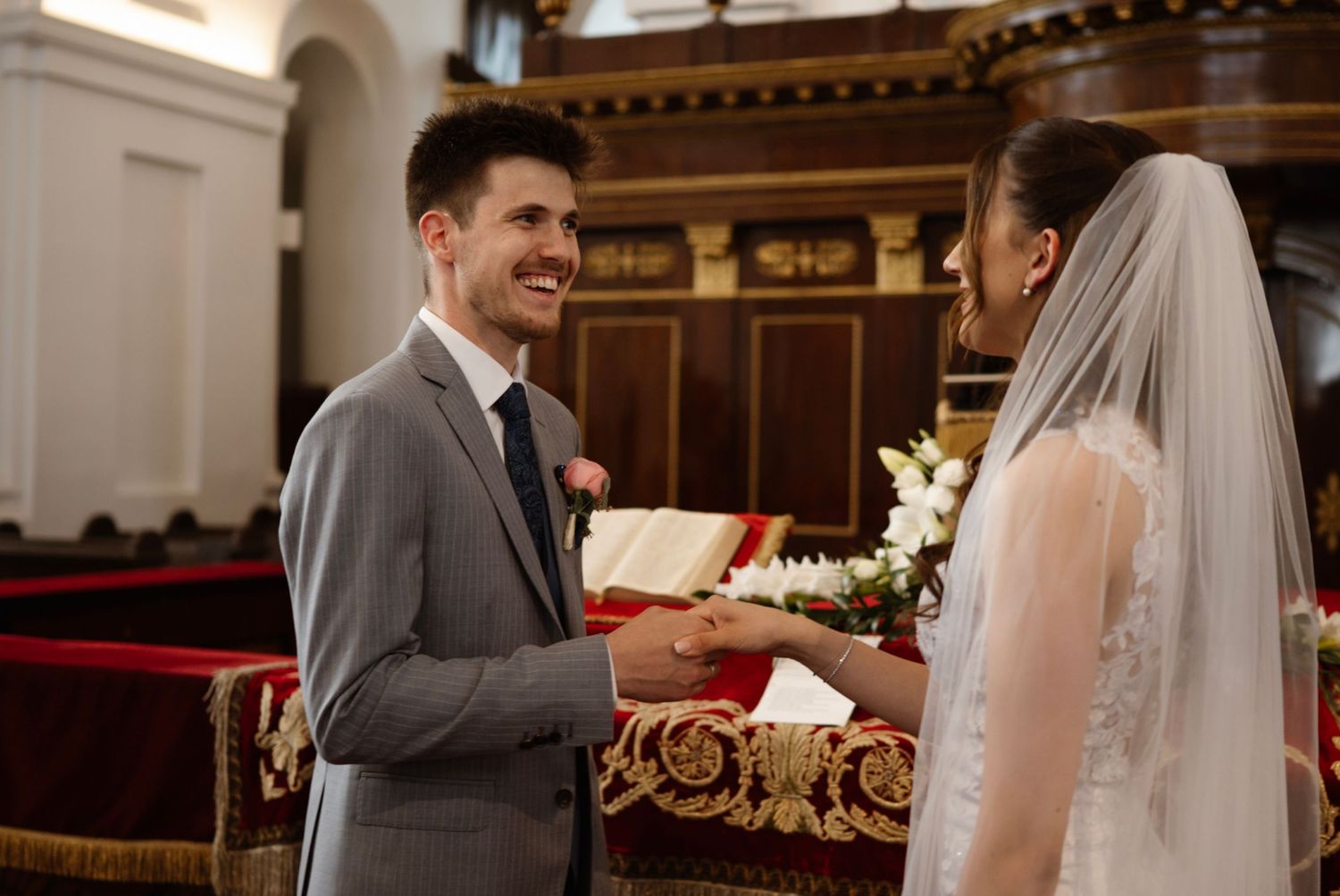 Groom in gray suit smiling at bride in white veil during wedding ceremony inside an elegant church.