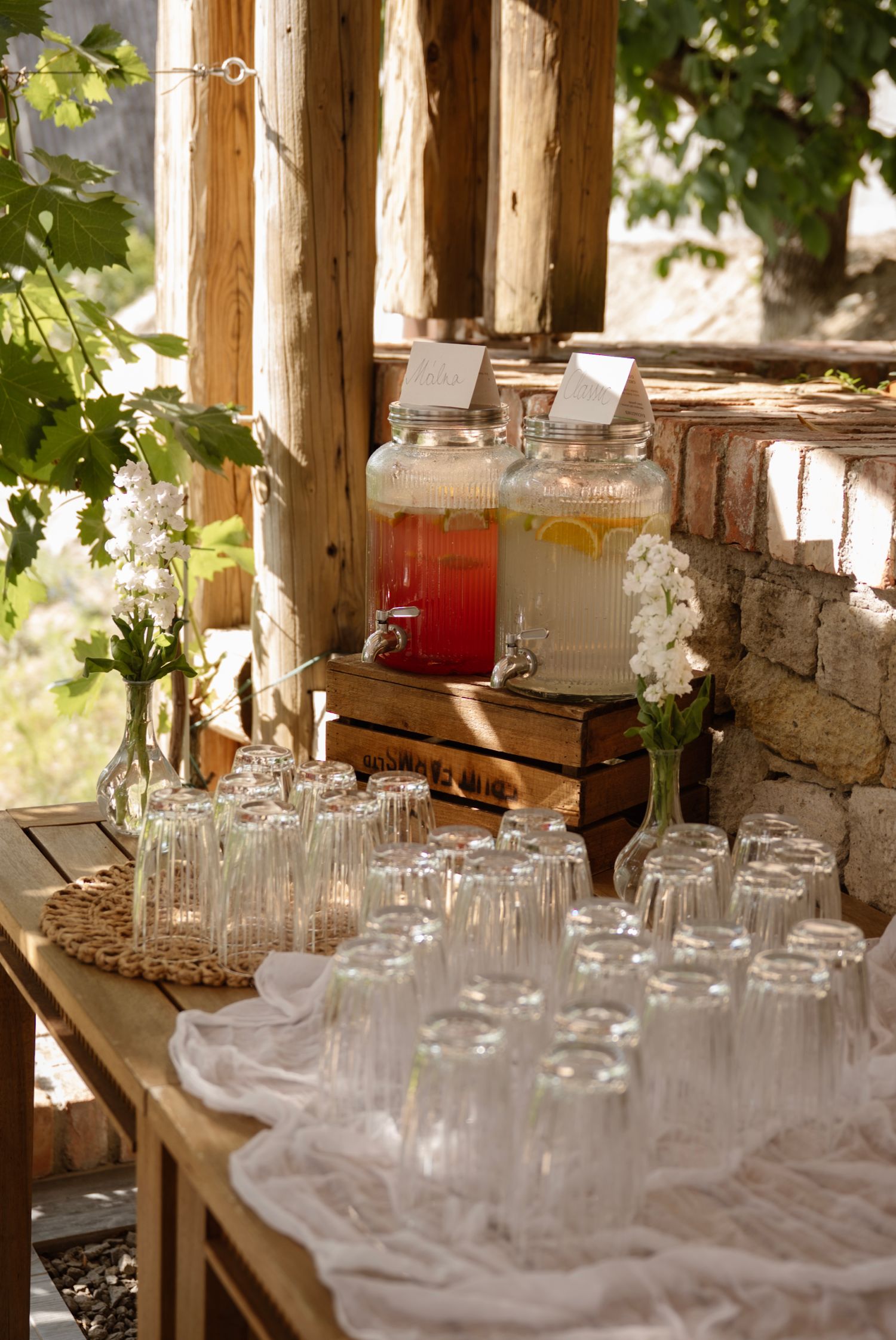 Rustic outdoor wedding drink station with mason jar dispensers of lemonade and pink punch surrounded by empty glasses.