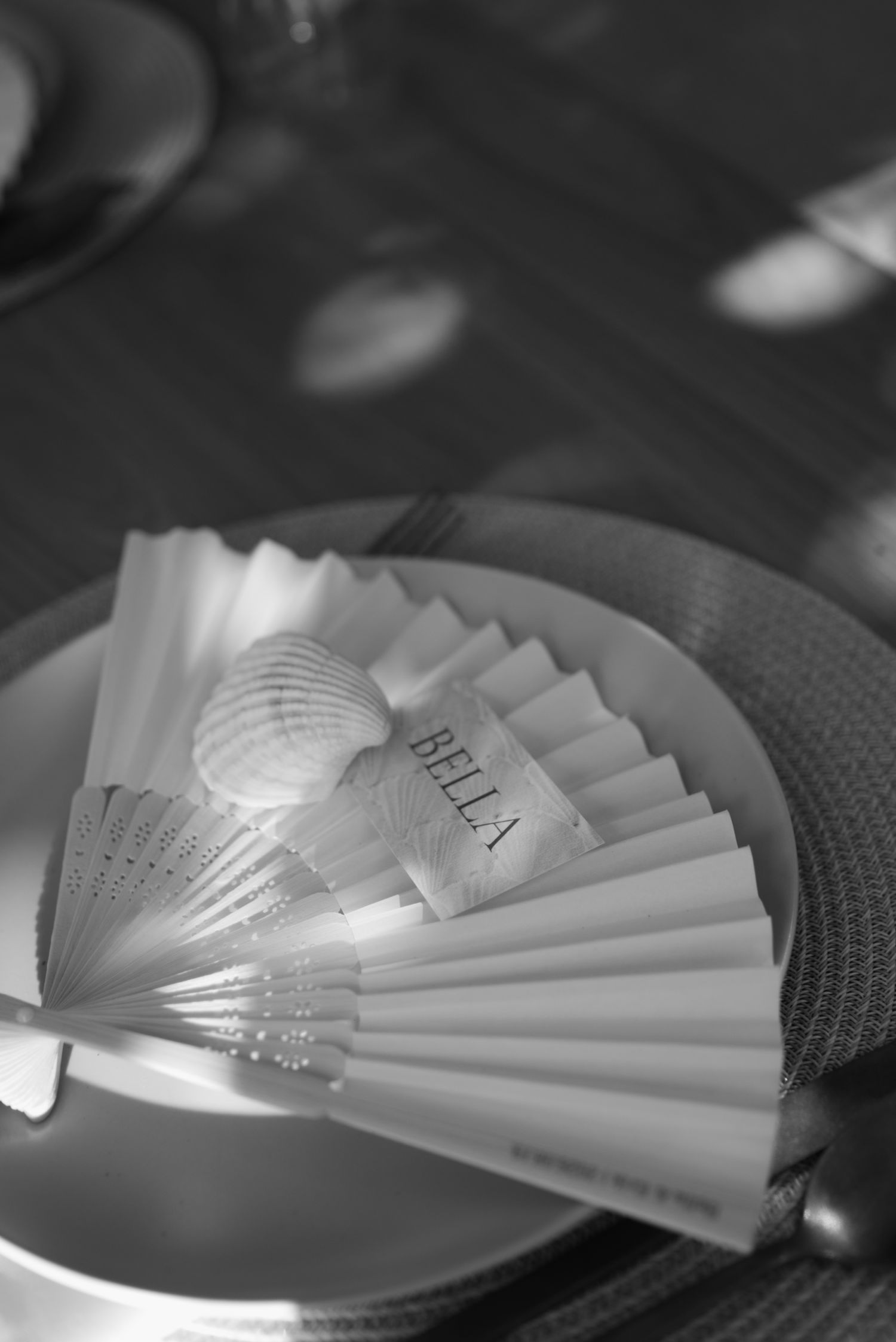 Black and white photo of folded paper fan place card reading Bella with seashell on wedding plate.