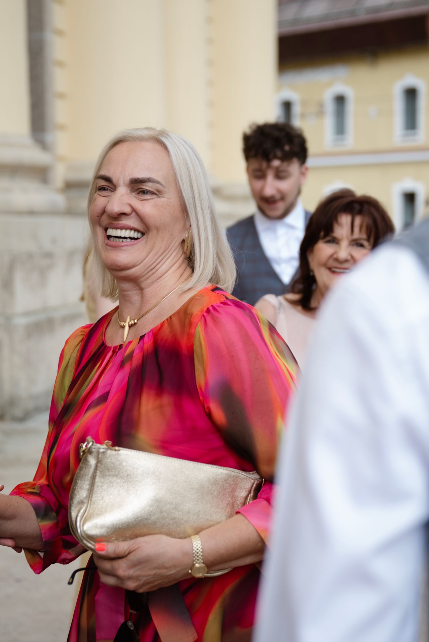 Smiling blonde woman in colorful pink dress holding gold clutch at outdoor wedding celebration.