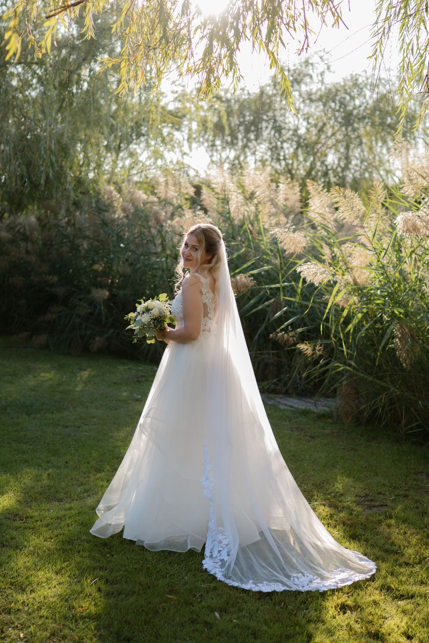 Bride in flowing white gown poses gracefully under willow trees in garden setting.