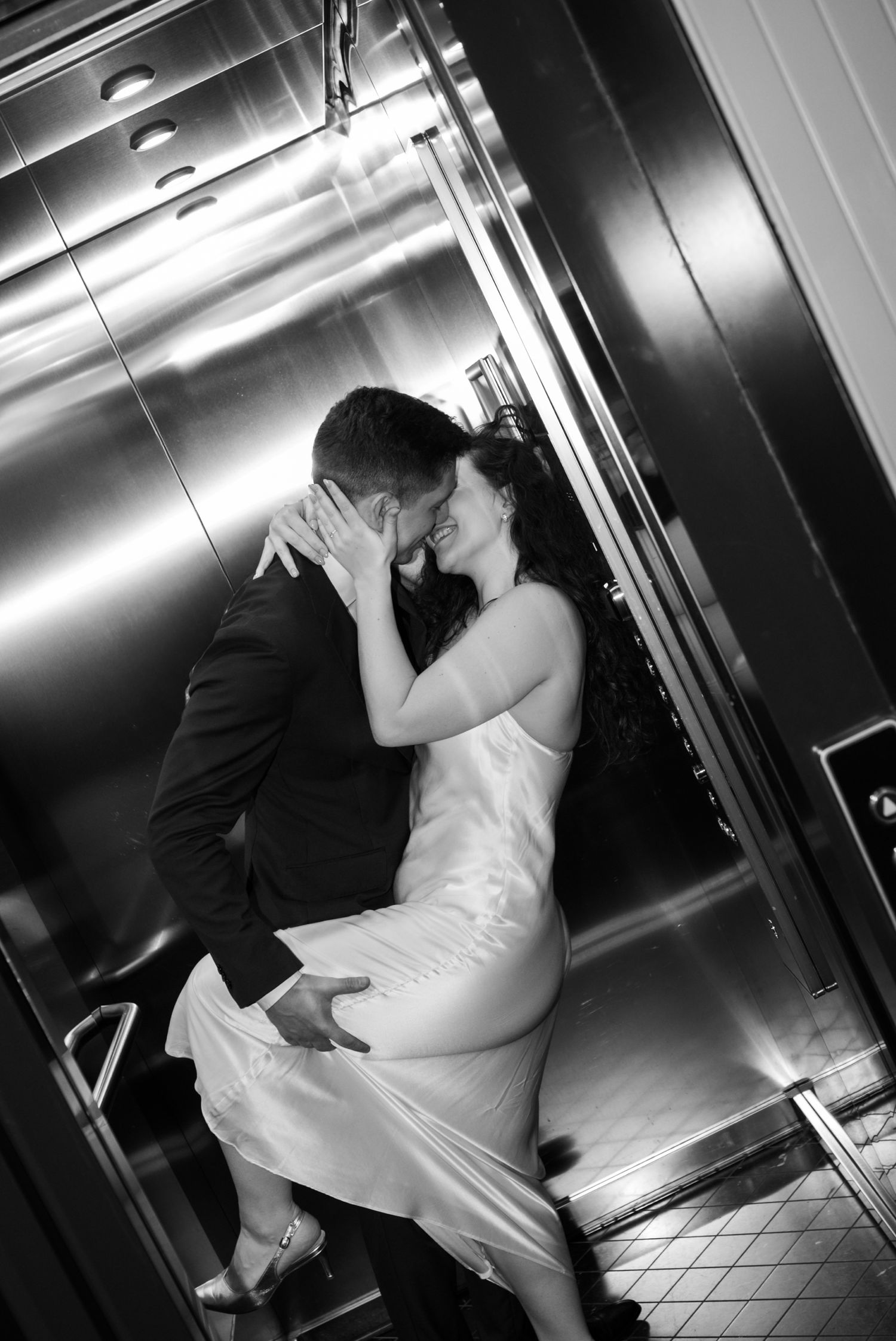 Black and white photo of a couple sharing a romantic kiss in a shiny elevator, bride in white dress.