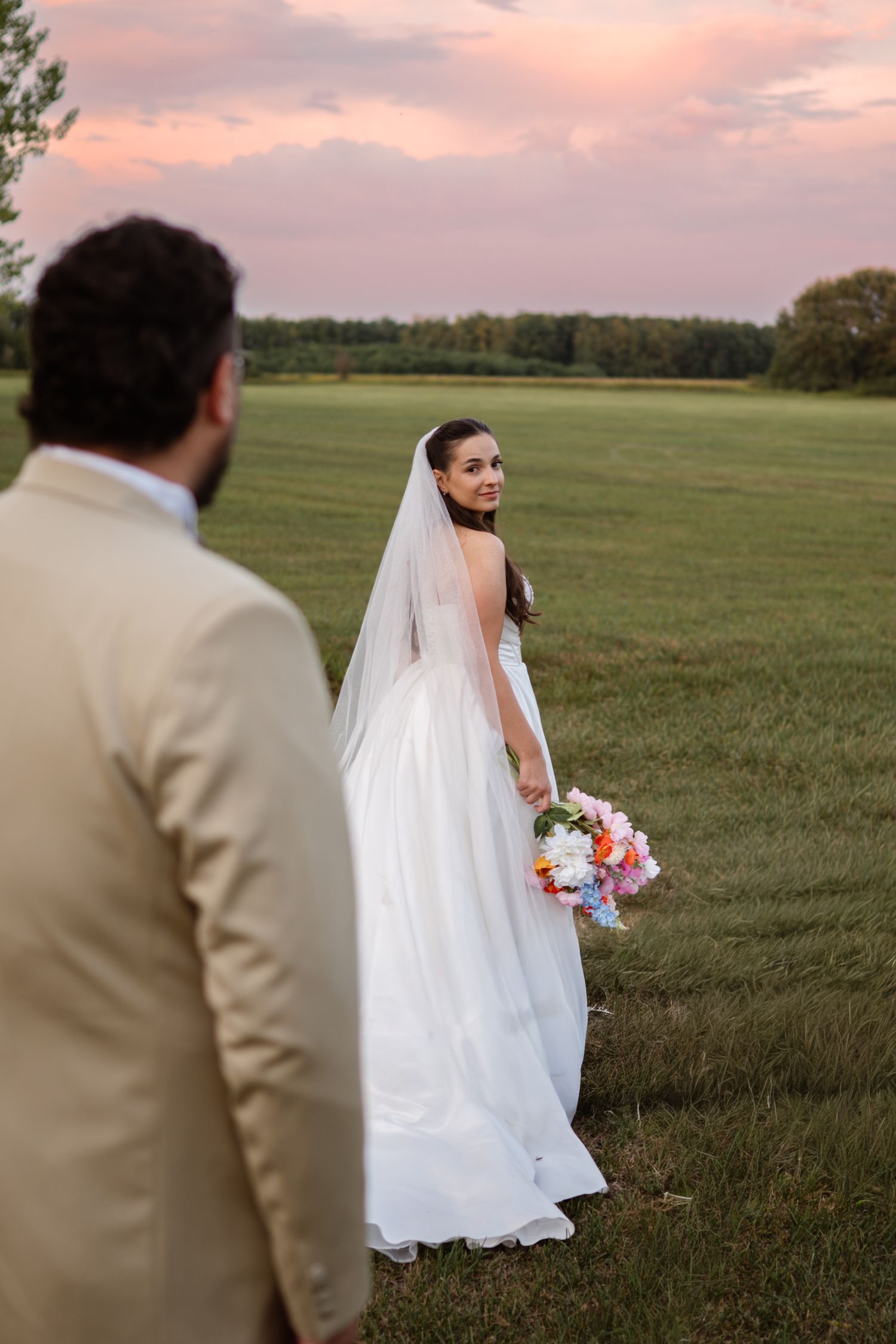 A bride in a white gown walking through a sunset-lit field.