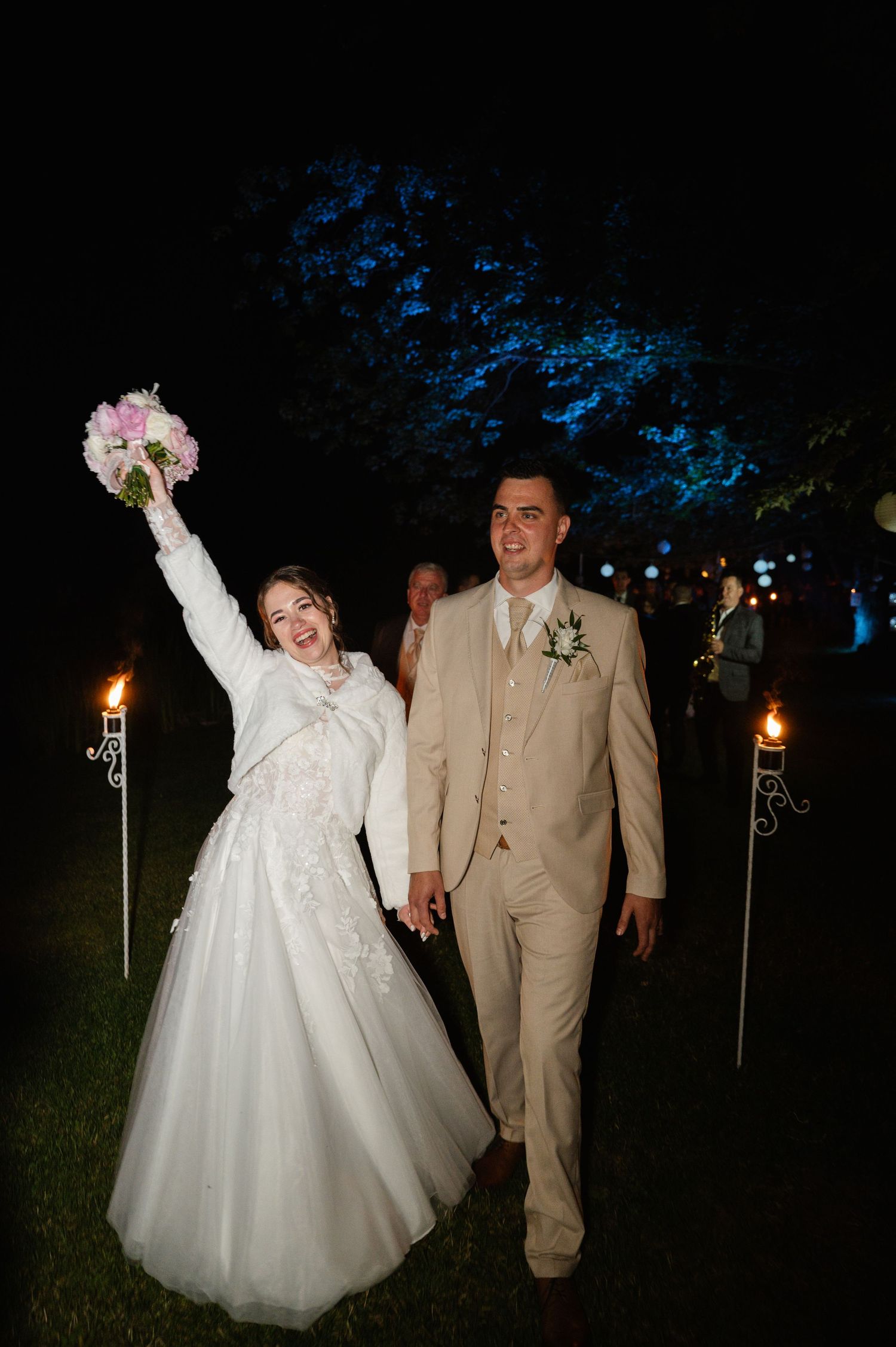 Newlyweds celebrate outdoors at night surrounded by tiki torches.