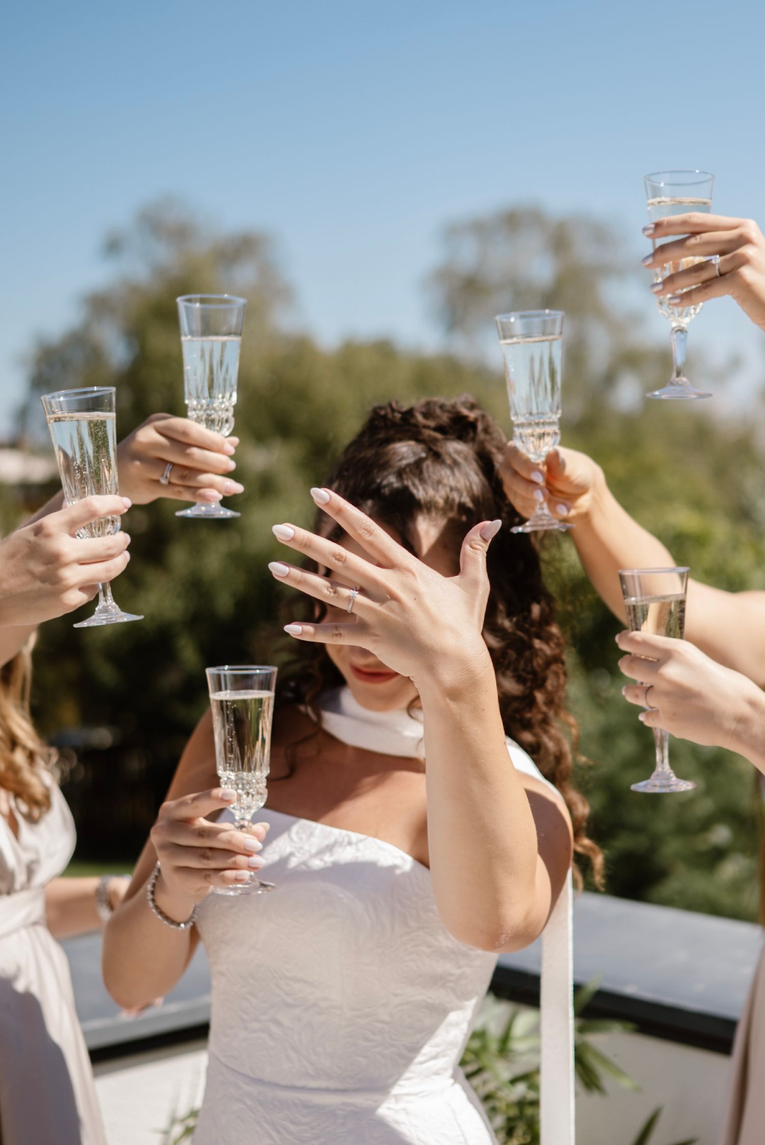 Bride in white dress raising hand among champagne toast with bridesmaids outdoors on sunny wedding day.