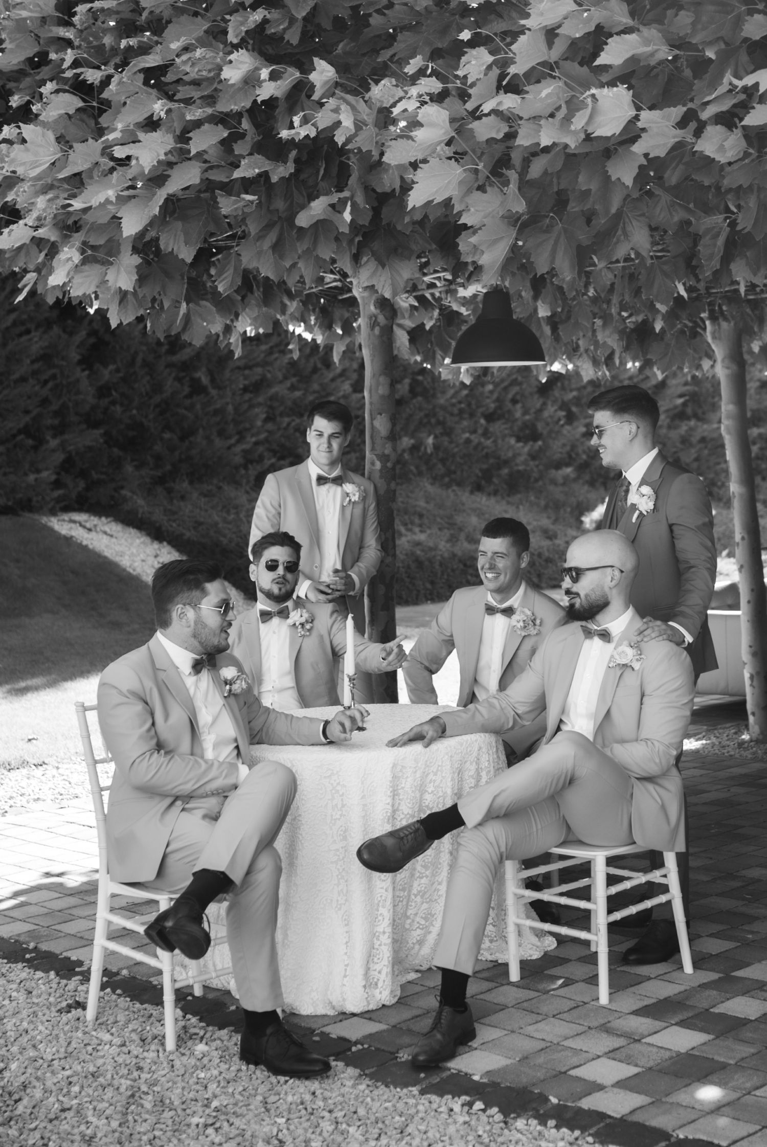 Black and white photo of groomsmen in bow ties and suits gathered around a table outdoors under trees at a wedding.
