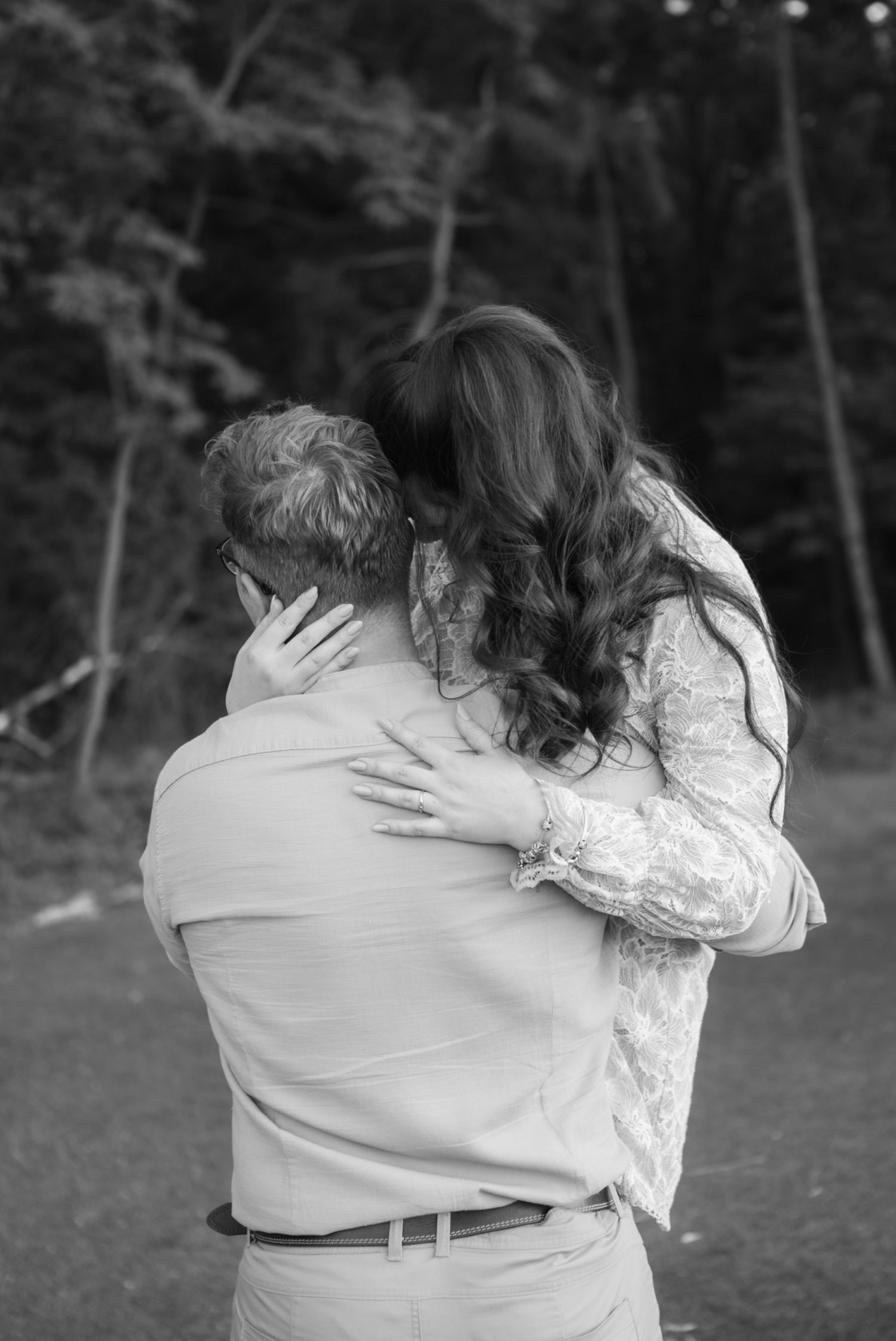Black and white photo of a couple embracing outdoors, woman with long wavy hair hugging man from behind near trees.