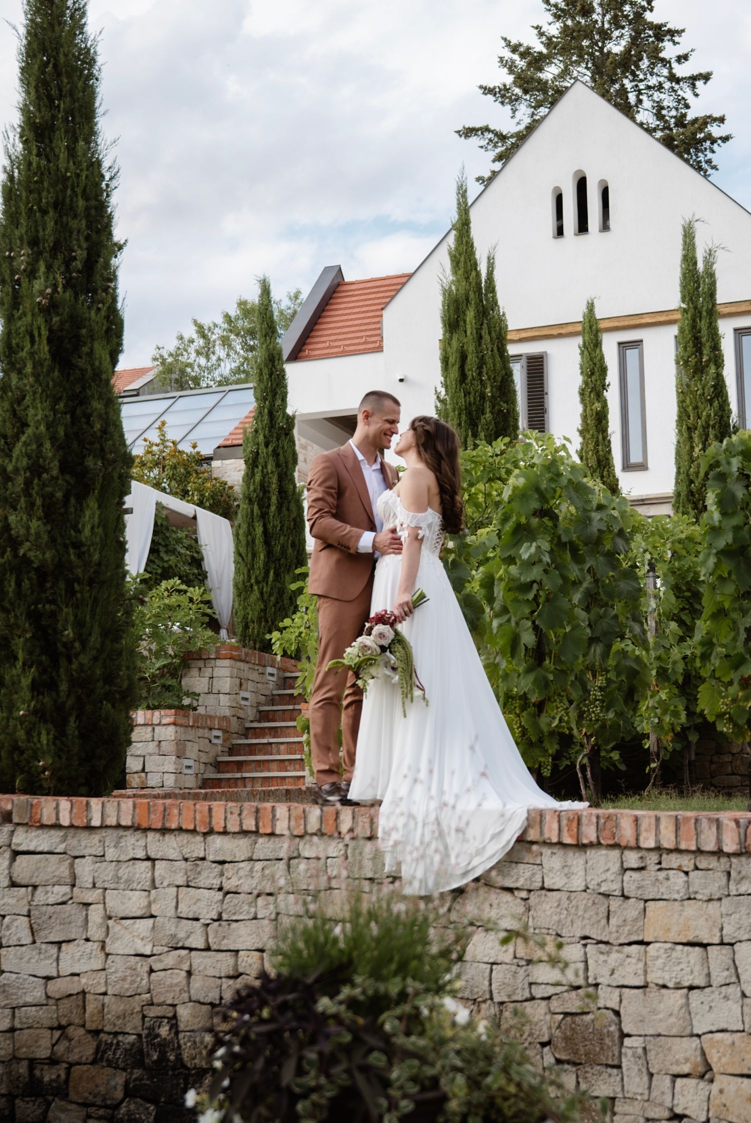 Bride and groom share romantic moment on stone terrace steps surrounded by cypress trees and greenery.