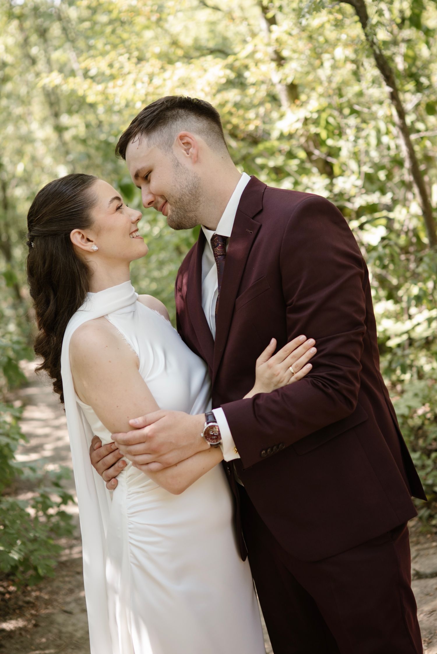 A bride in white gown and groom in burgundy suit share a romantic moment in a lush green forest setting.