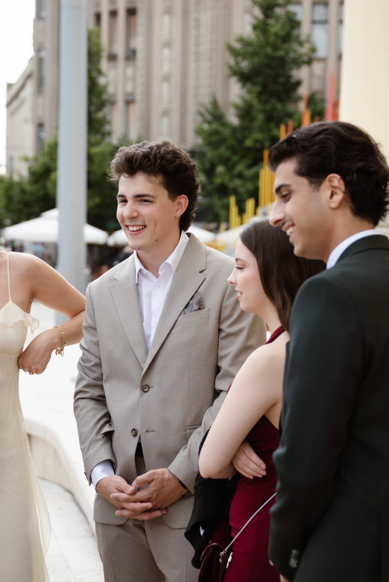 Young adults in formal attire socializing outdoors at an elegant event, with a smiling man in a gray suit.