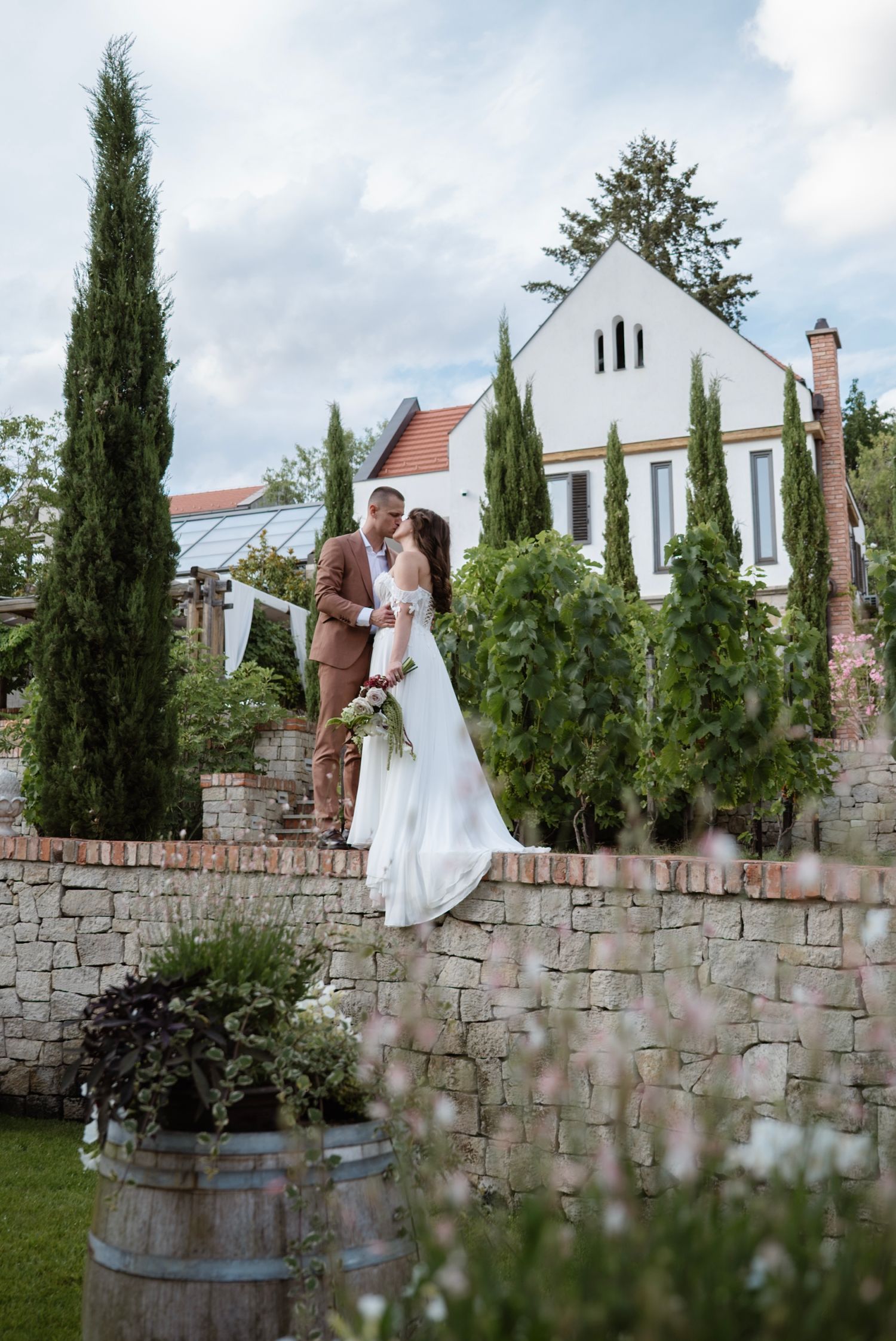 Bride and groom sharing a romantic moment on a stone wall surrounded by cypress trees and a white house backdrop.