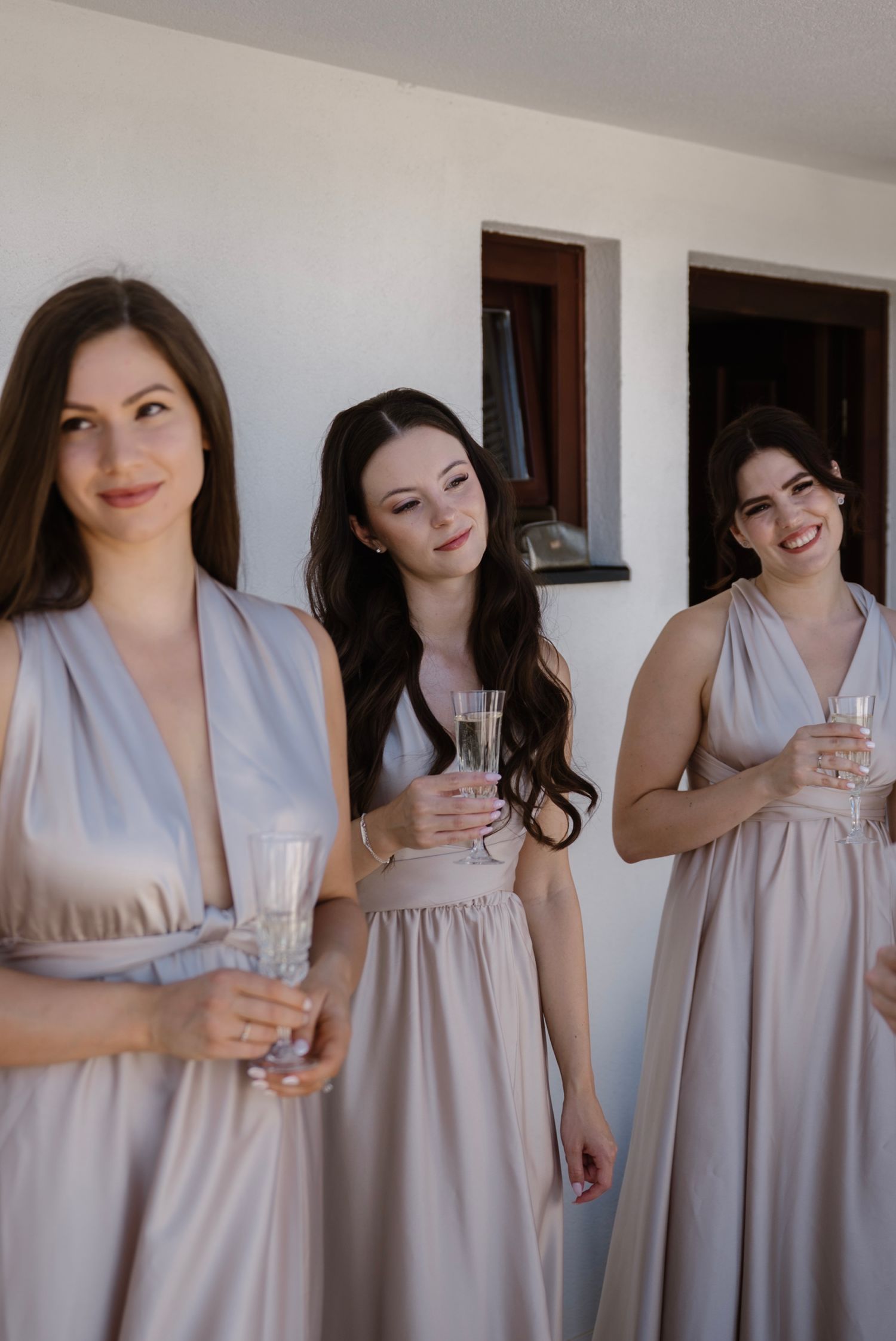Three bridesmaids in elegant taupe satin gowns holding champagne flutes, smiling during wedding preparations indoors.