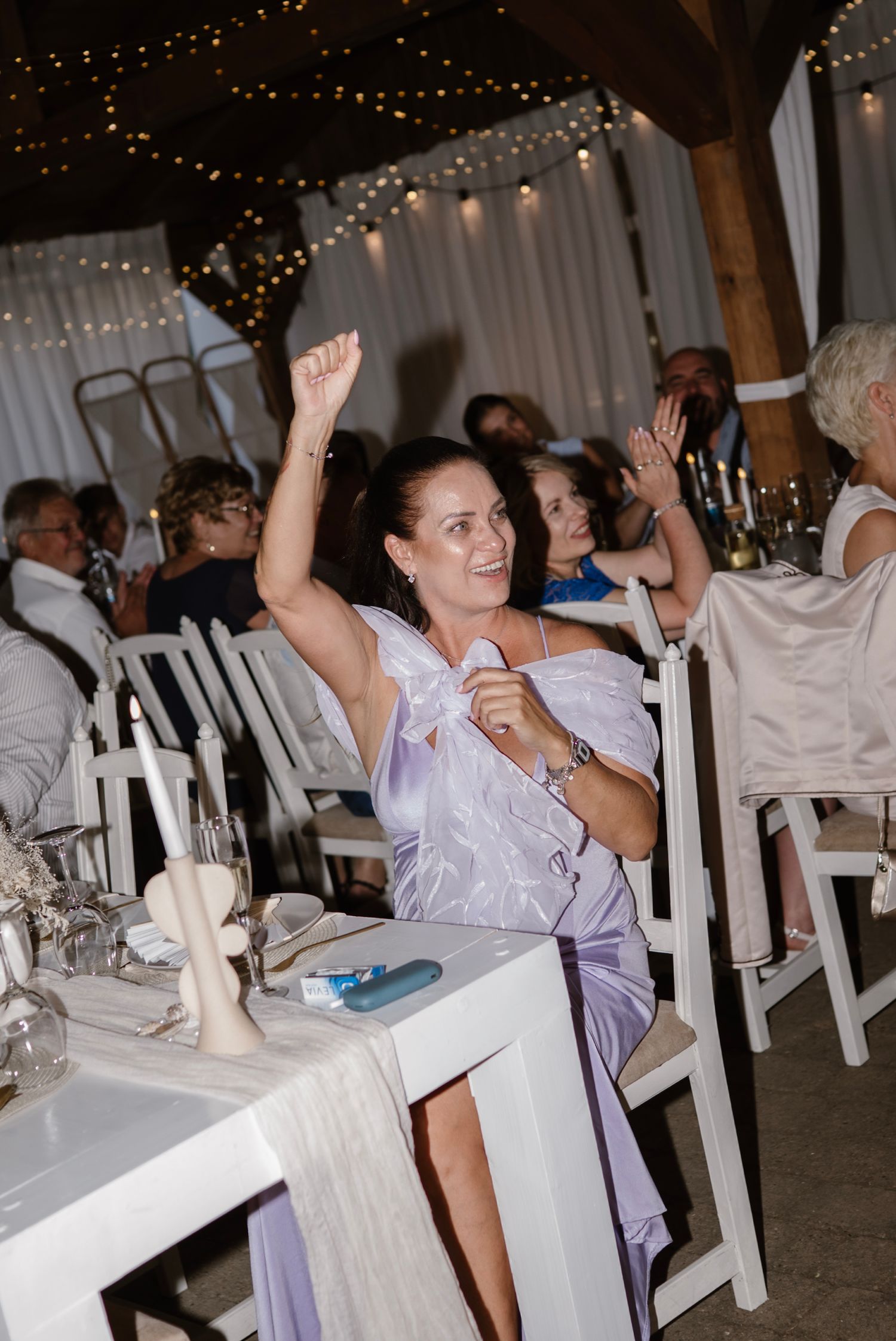 Woman in lavender dress raises fist celebrating at elegant wedding reception with fairy lights.