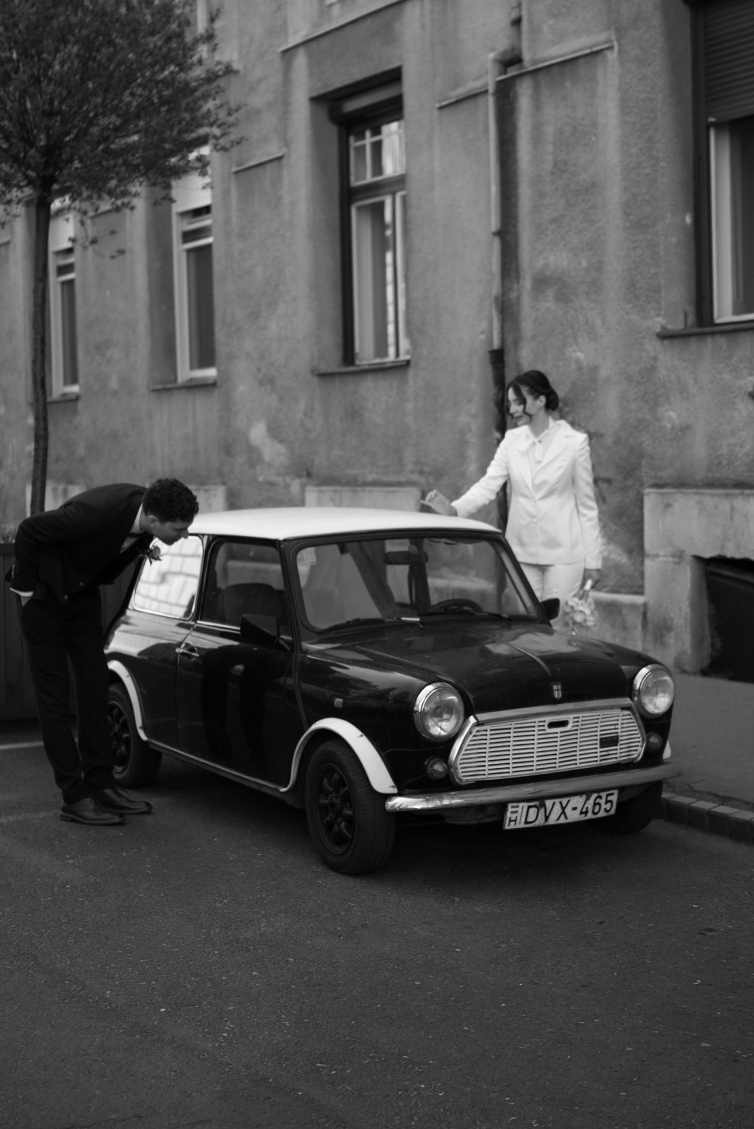 Black and white photo of a couple with a classic Mini Cooper car on a street beside a European building.