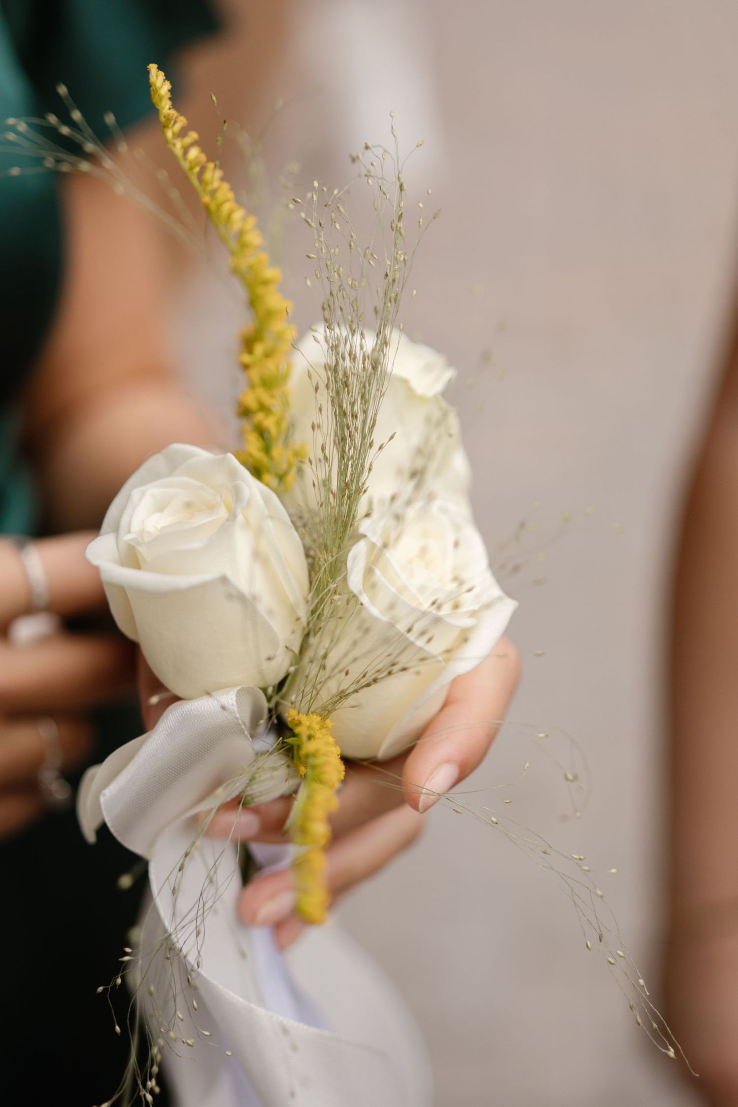 A delicate white rose and yellow flower boutonniere held in someone's hand.