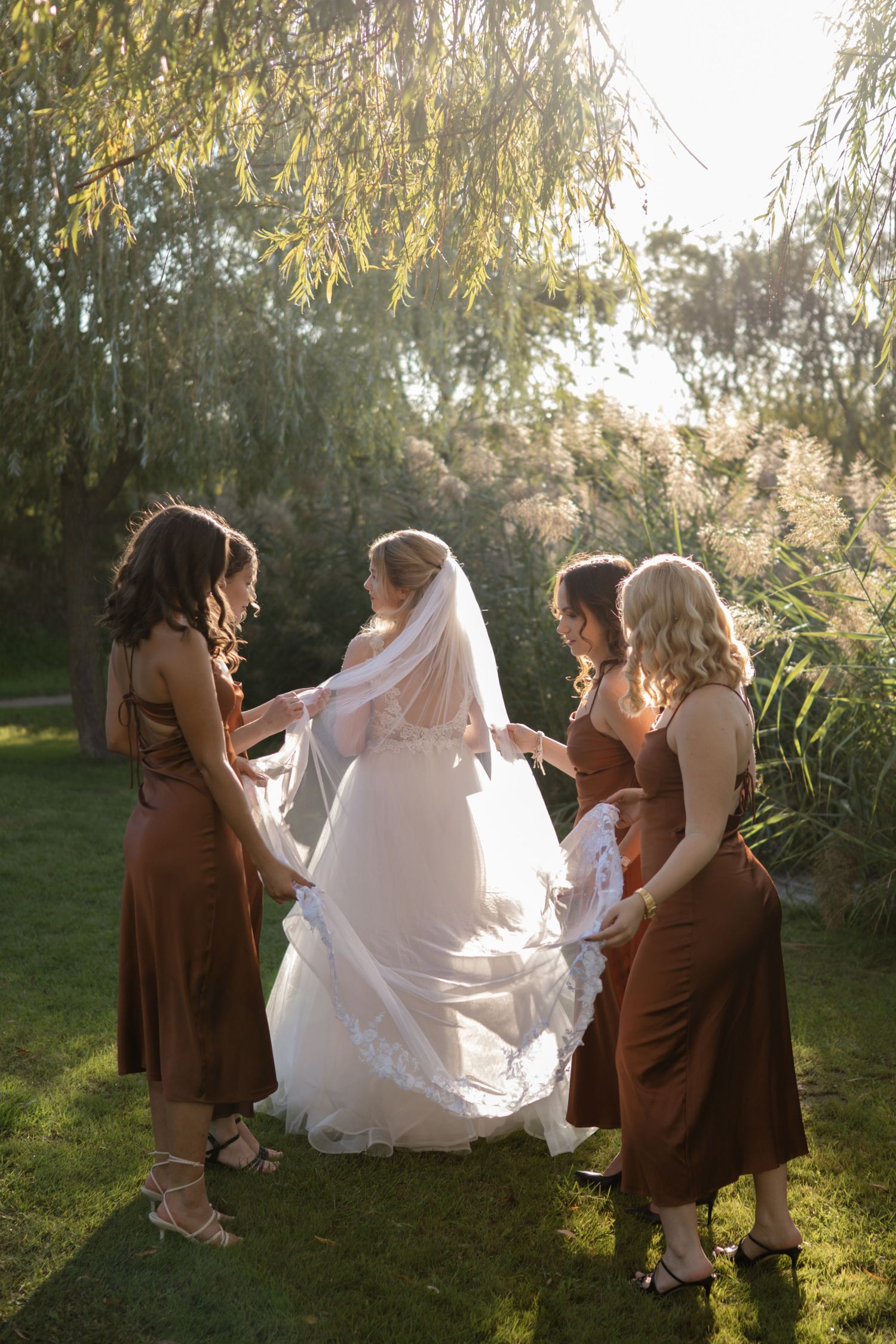 Bridesmaids in brown dresses help arrange bride's wedding gown in golden sunlight.