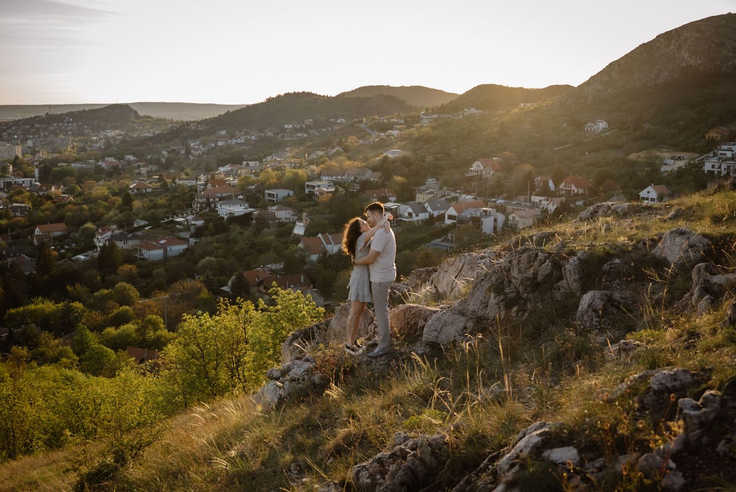 Couple embracing on rocky hilltop at golden sunset with scenic village and green hills in background.