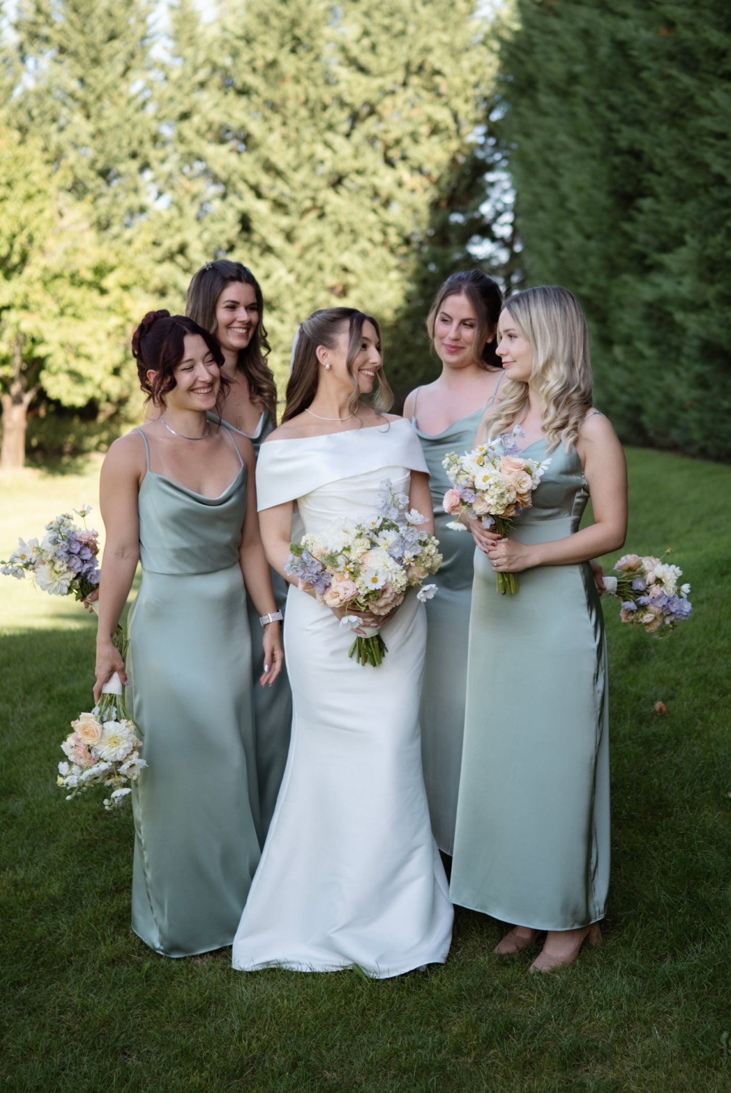 Bride in white off-shoulder gown with four bridesmaids in sage green dresses holding floral bouquets outdoors.