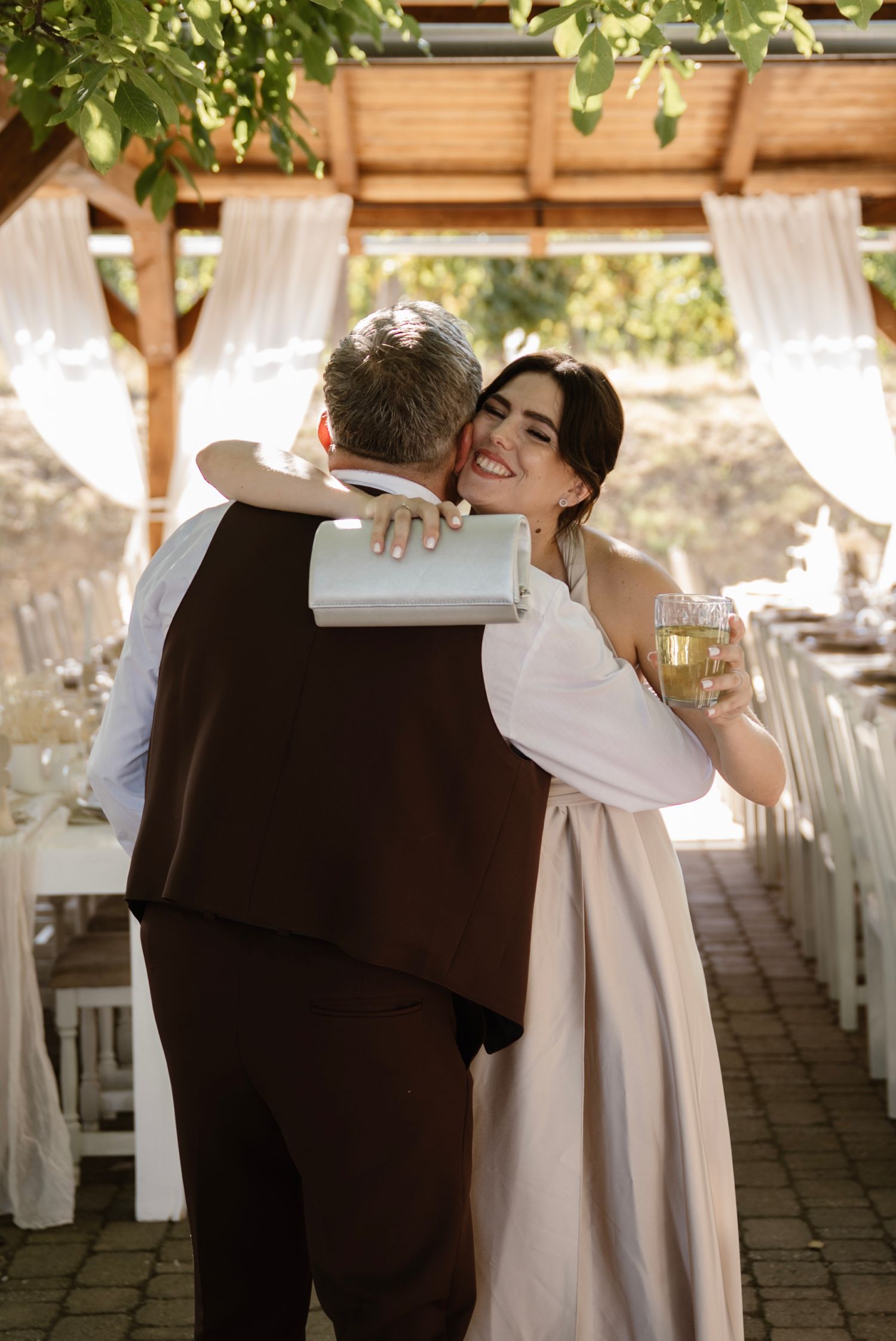A smiling bride in a white dress hugs a groom in a brown suit, holding champagne at an outdoor wedding reception.