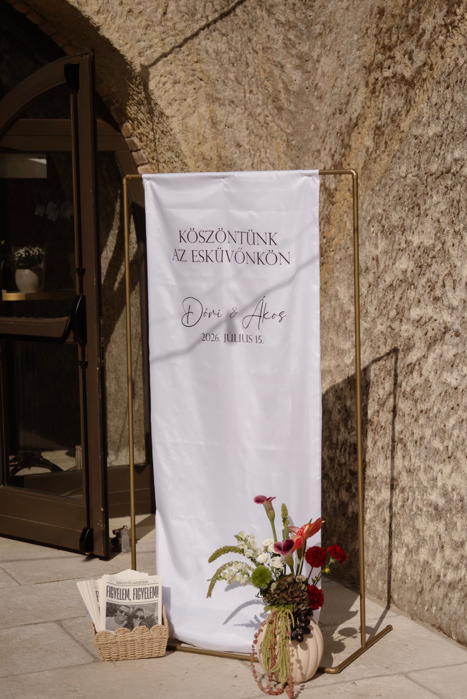 Elegant wedding welcome sign in Hungarian on a gold frame stand with floral arrangement outside a stone venue.