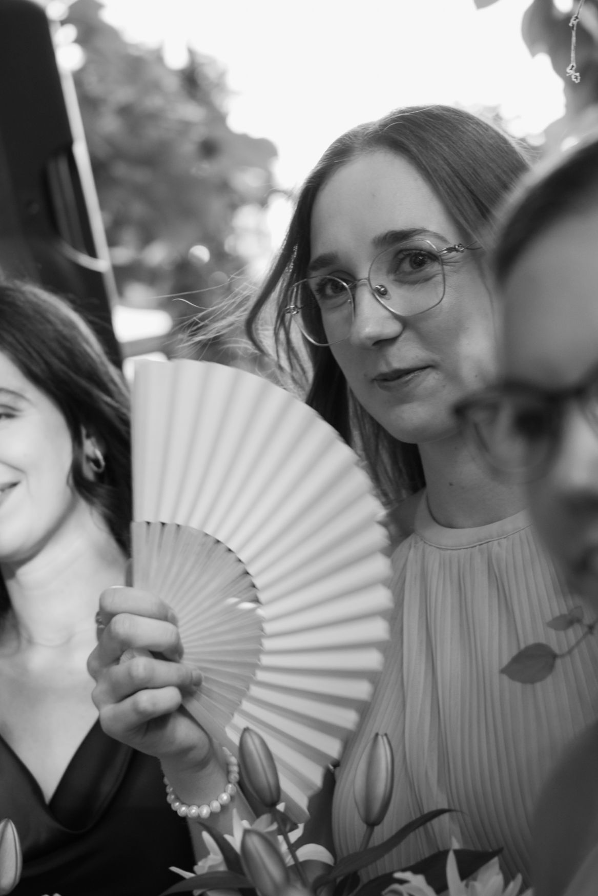 Black and white photo of a woman wearing glasses holding a decorative hand fan at an outdoor event.