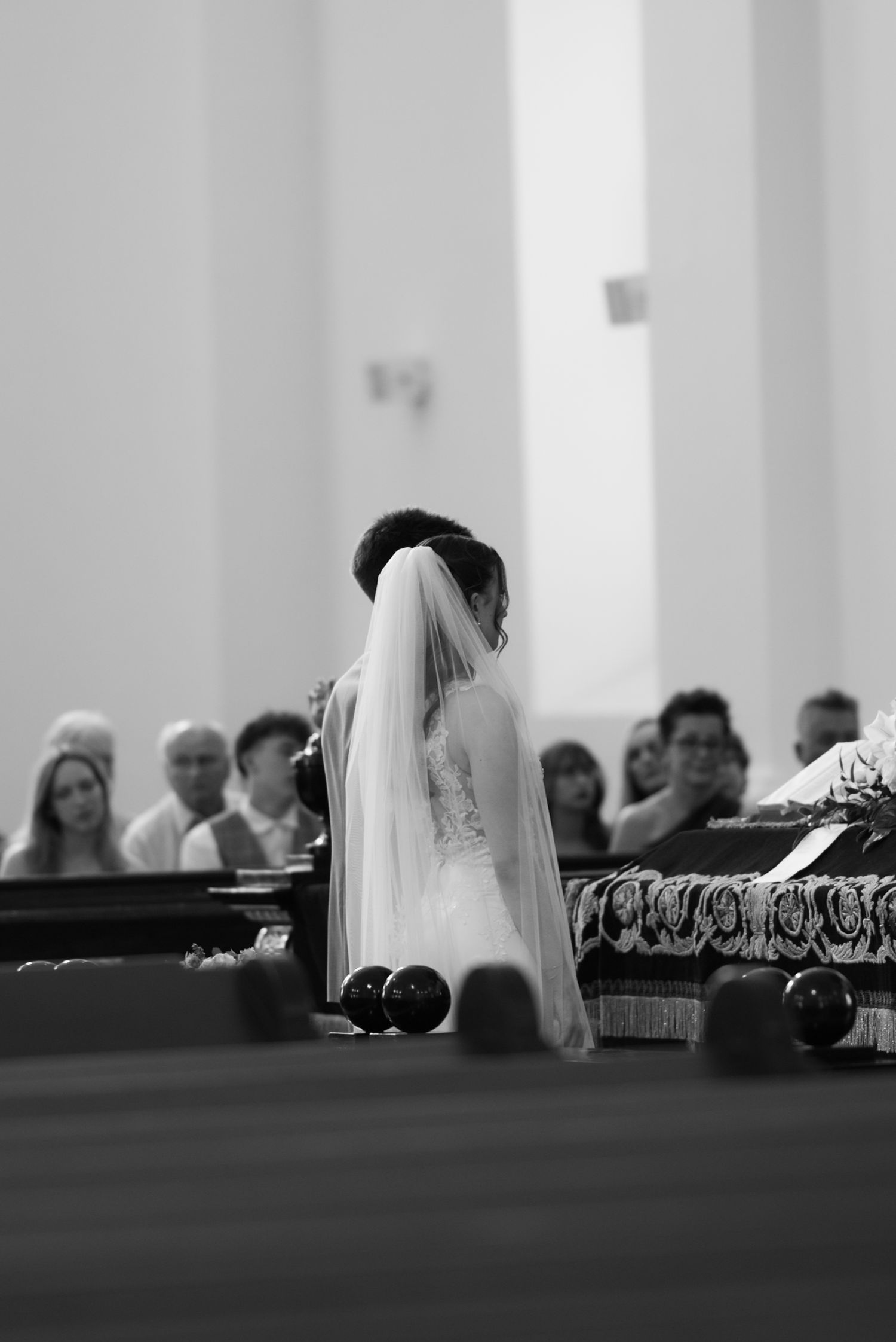 Bride in white wedding gown and veil kneeling at church altar during ceremony with guests seated in background.