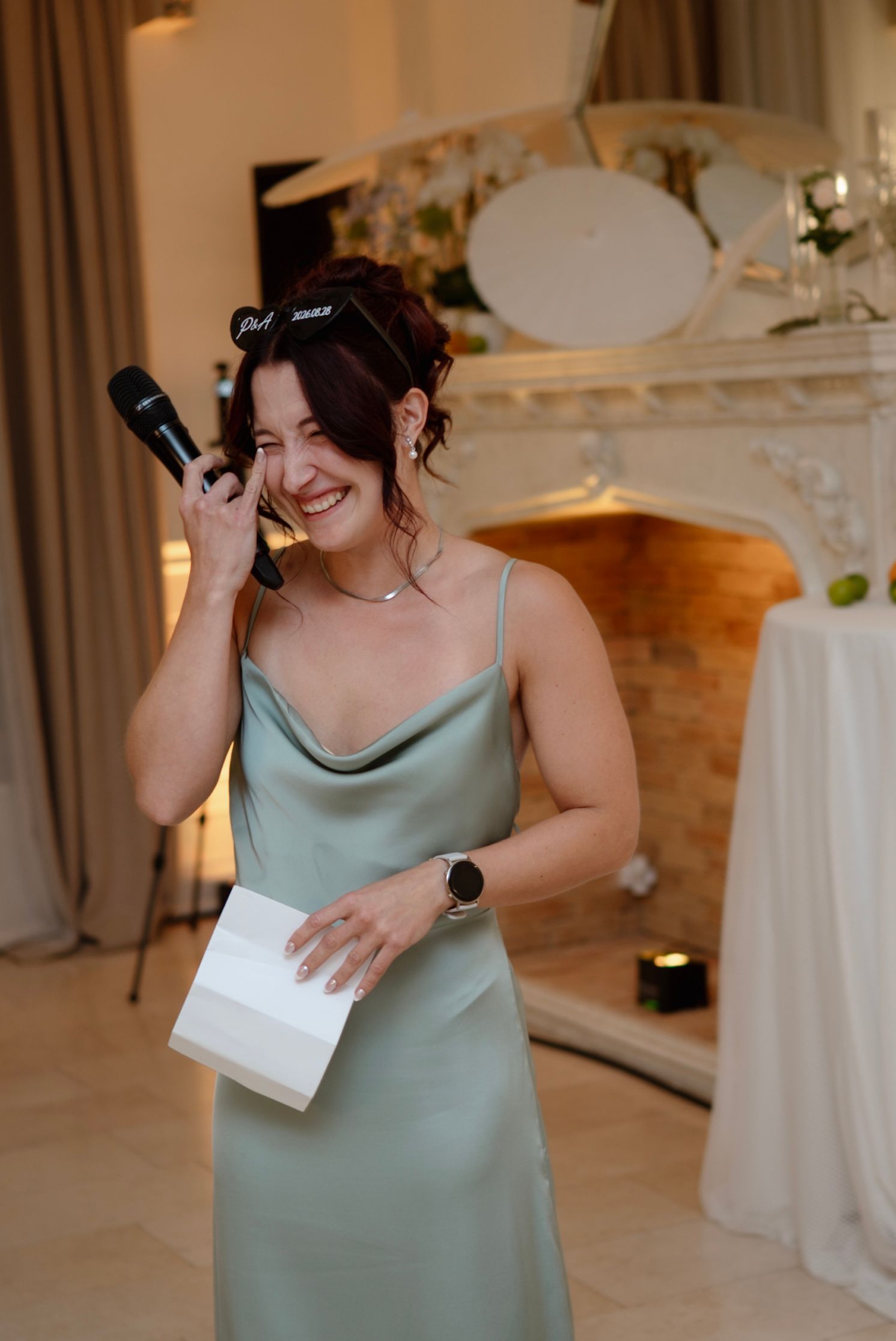 Woman in sage green satin dress giving a speech at a wedding reception, holding microphone and notes.