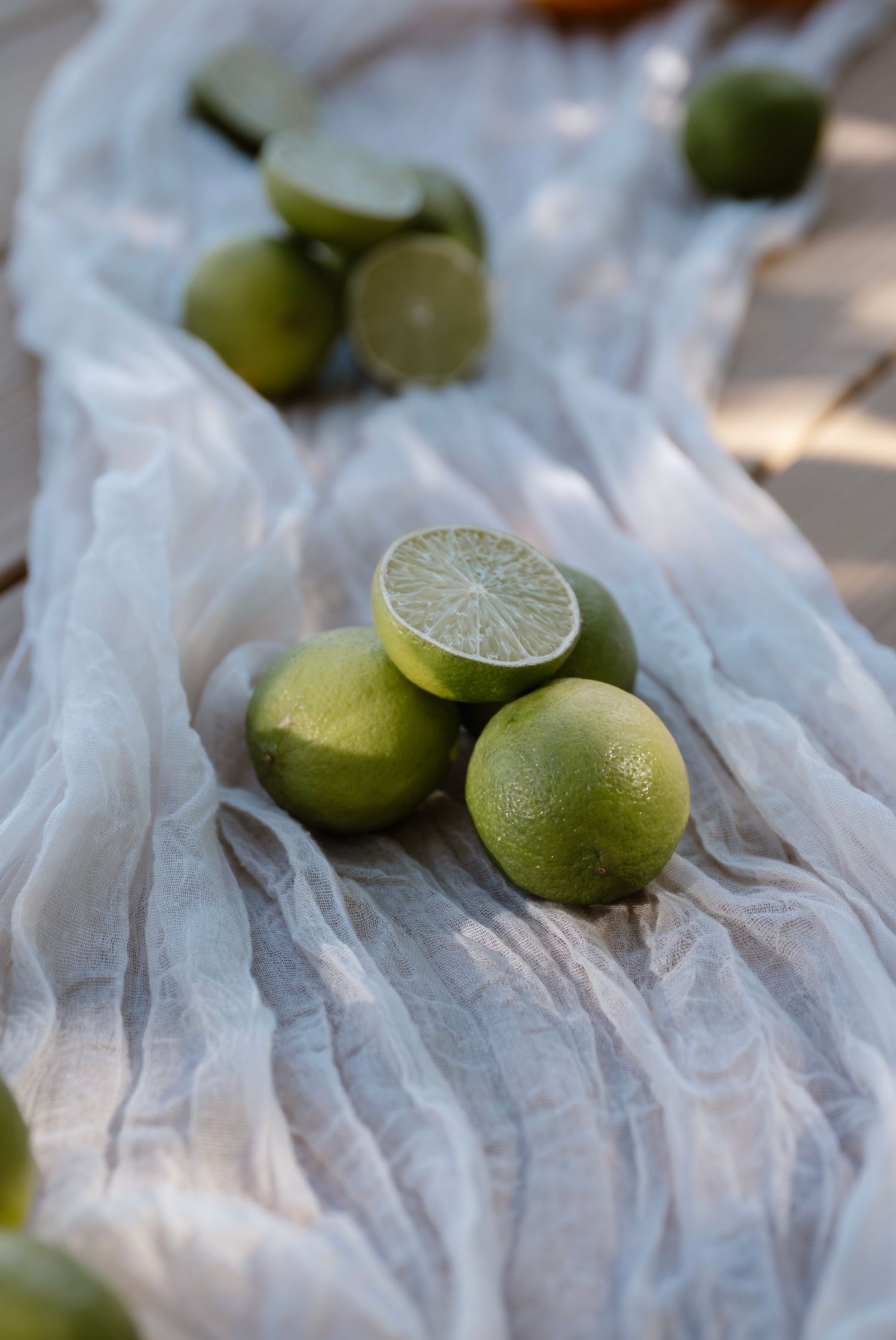Fresh green limes on white gauze fabric, one halved to reveal juicy interior, scattered arrangement in natural light.