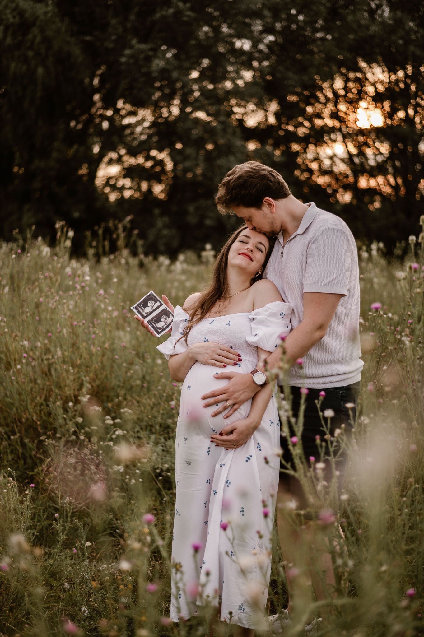 Expectant couple holding ultrasound photo in wildflower meadow at sunset.