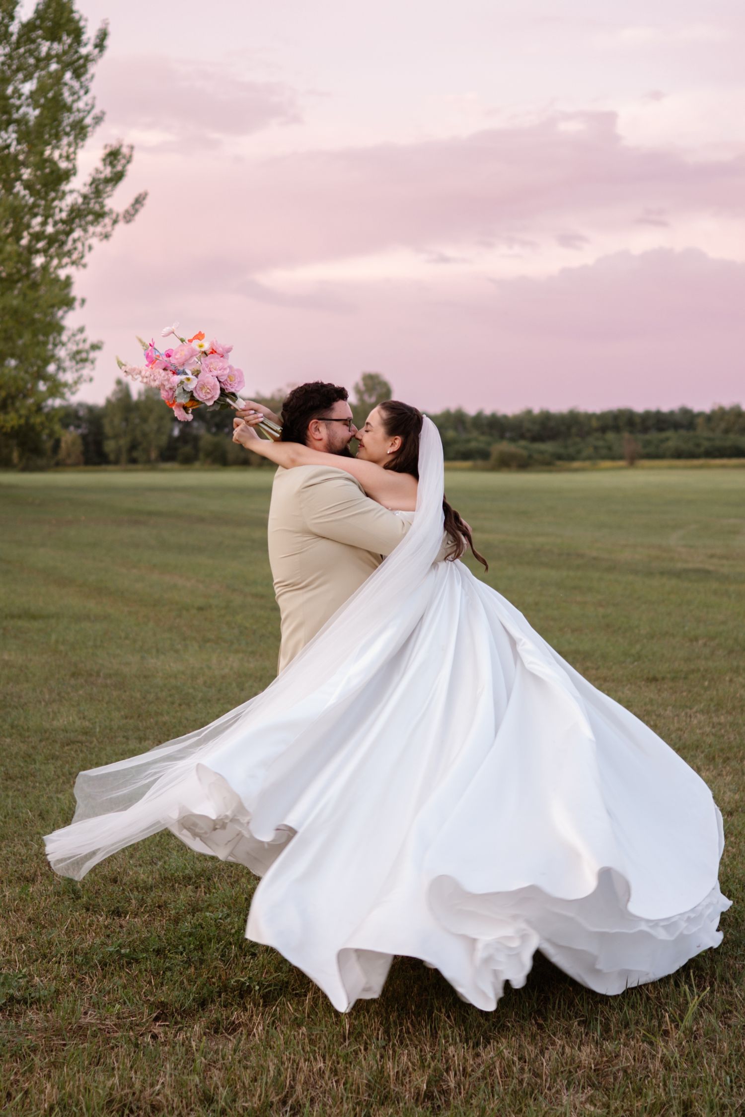 A romantic wedding couple dancing in a field at sunset with the bride's white dress flowing in the wind.