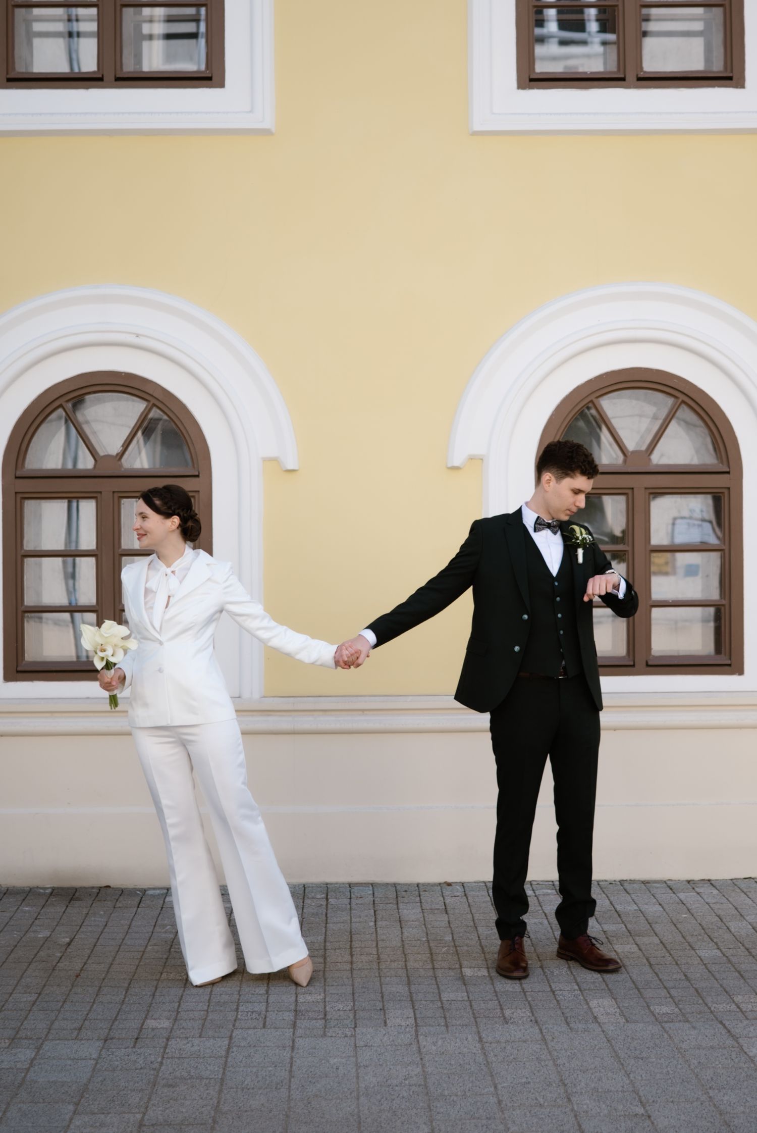 Bride in white pantsuit and groom in black tuxedo hold hands outside a yellow building with arched windows.