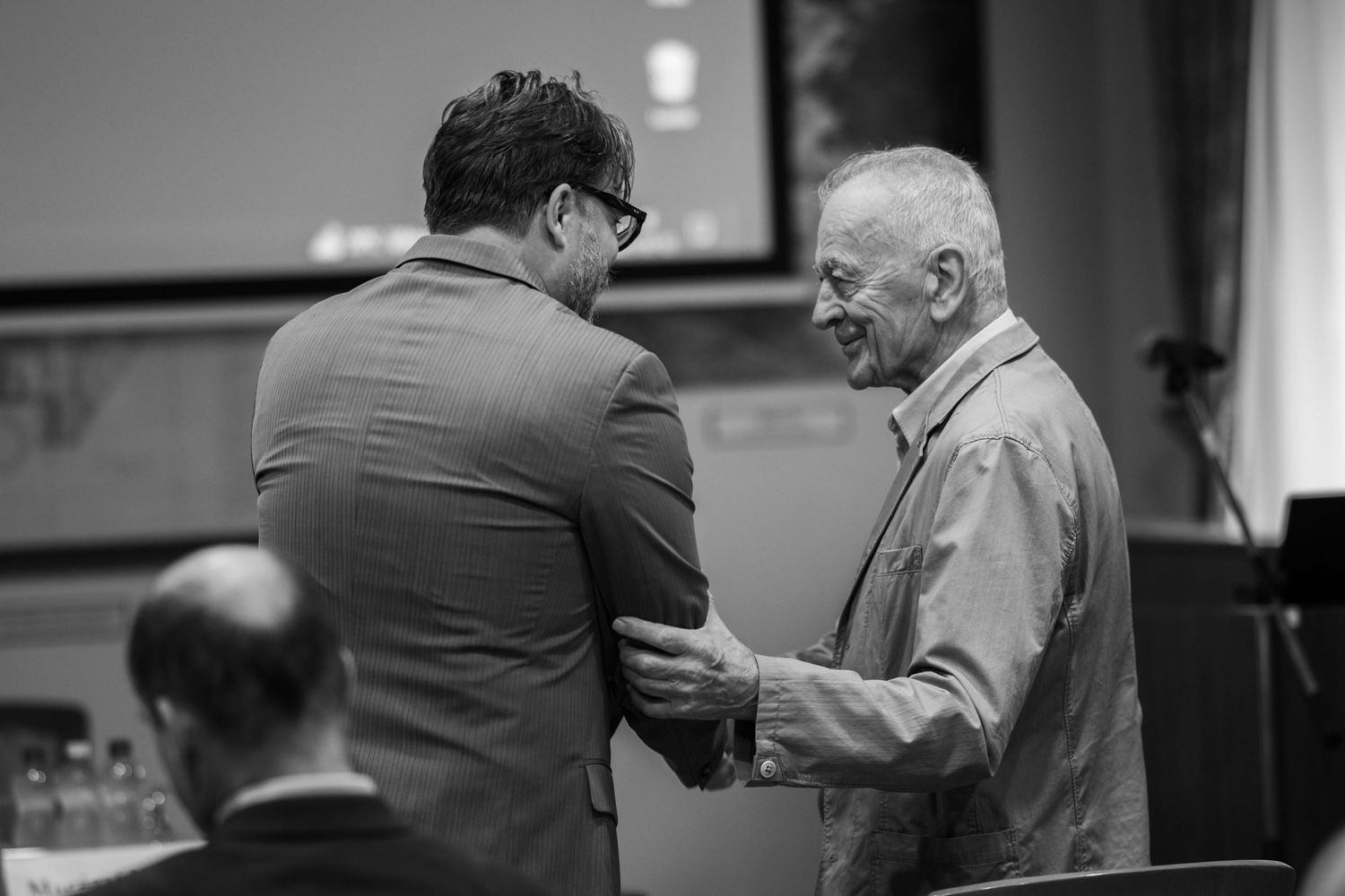 Two men in conversation at an indoor event, with one wearing glasses and a suit and the other in casual attire.