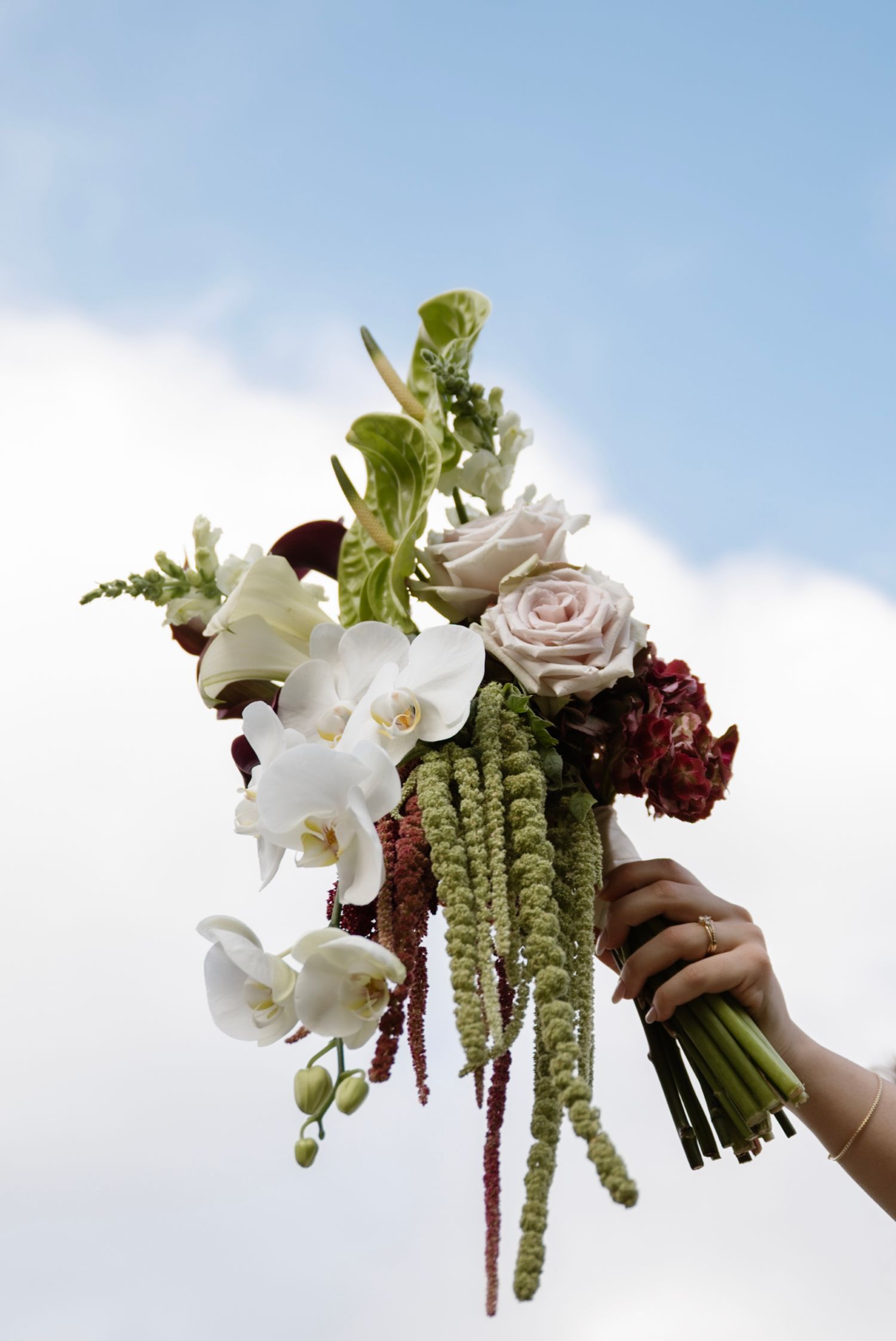 Hand holding a bridal bouquet with white orchids, pink roses, and burgundy flowers against a blue sky.