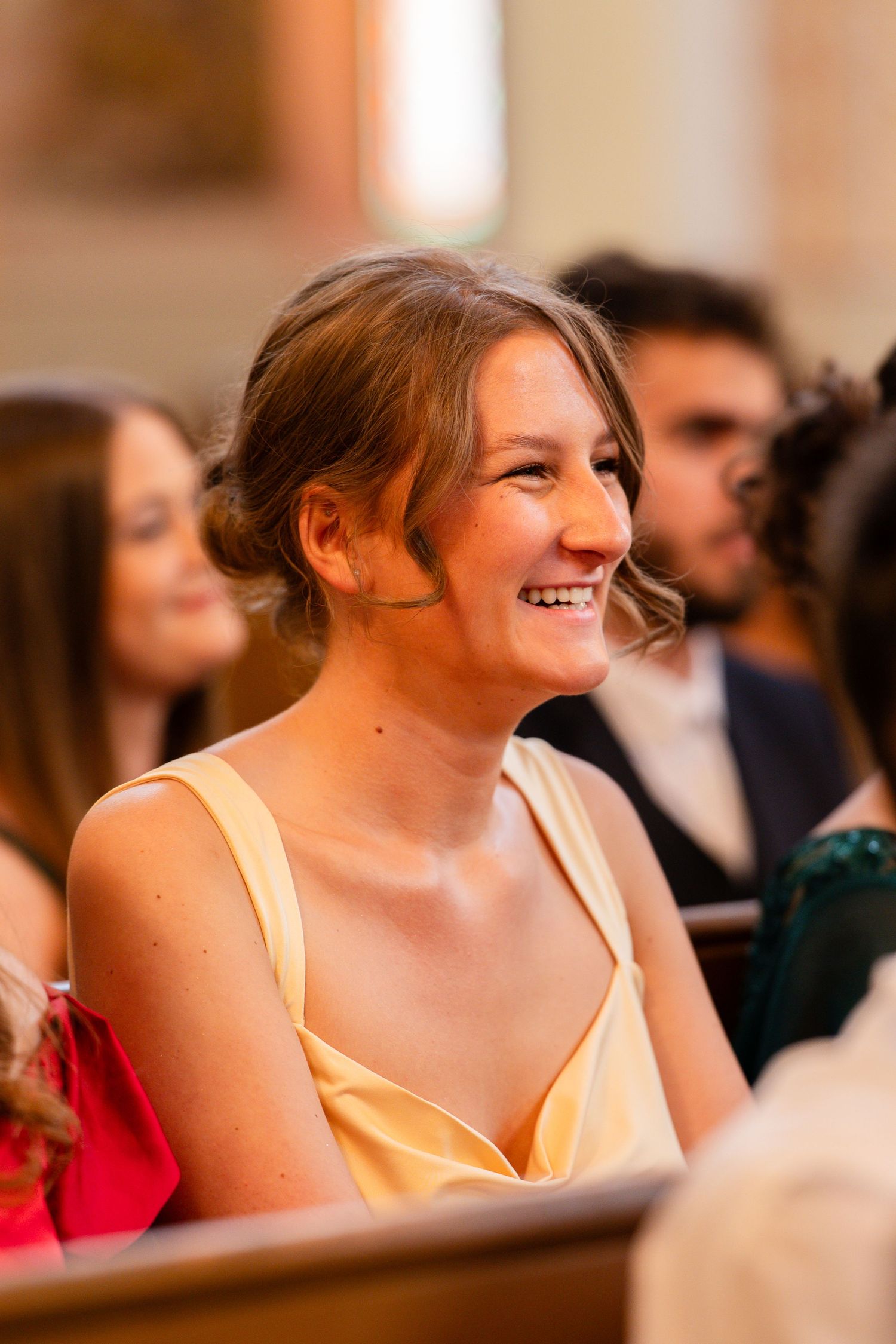 Wedding guest in a pale yellow dress smiles joyfully while seated in a church pew.