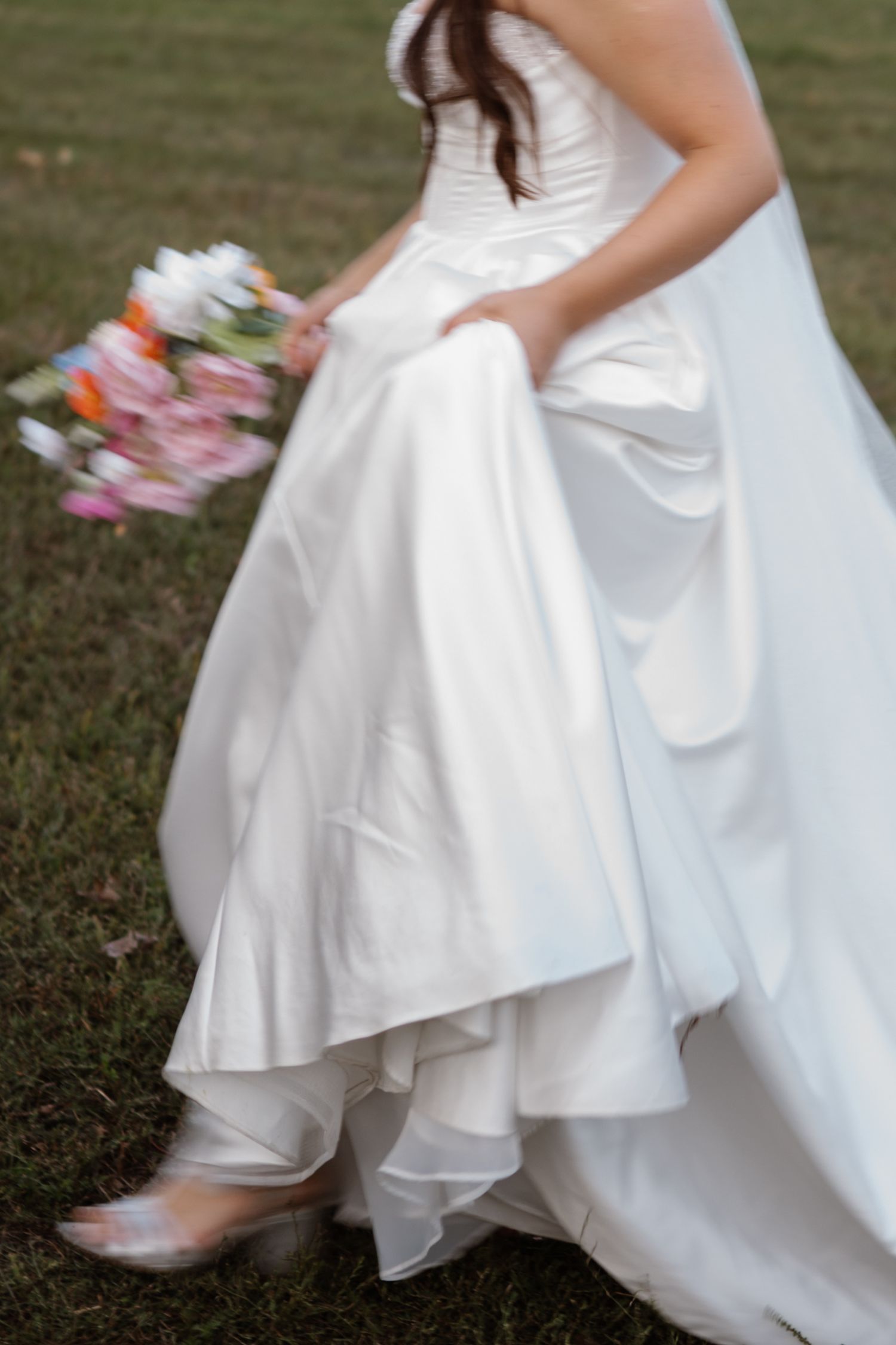 Close-up detail of a flowing white wedding dress and bouquet against grass.