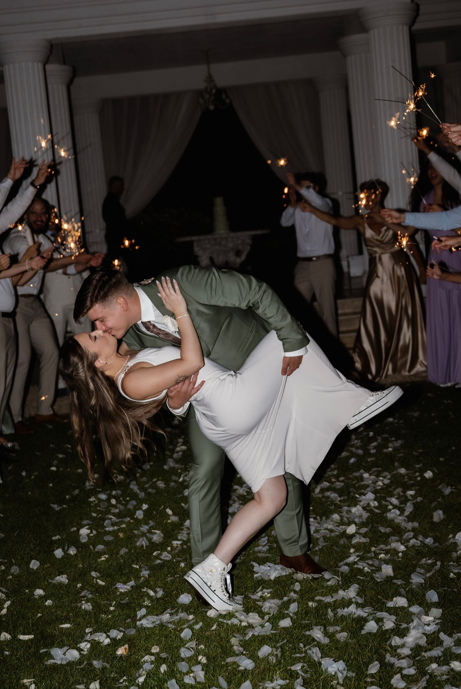 Bride and groom share a romantic dip kiss surrounded by sparkler-holding guests on a flower petal-covered lawn at night.