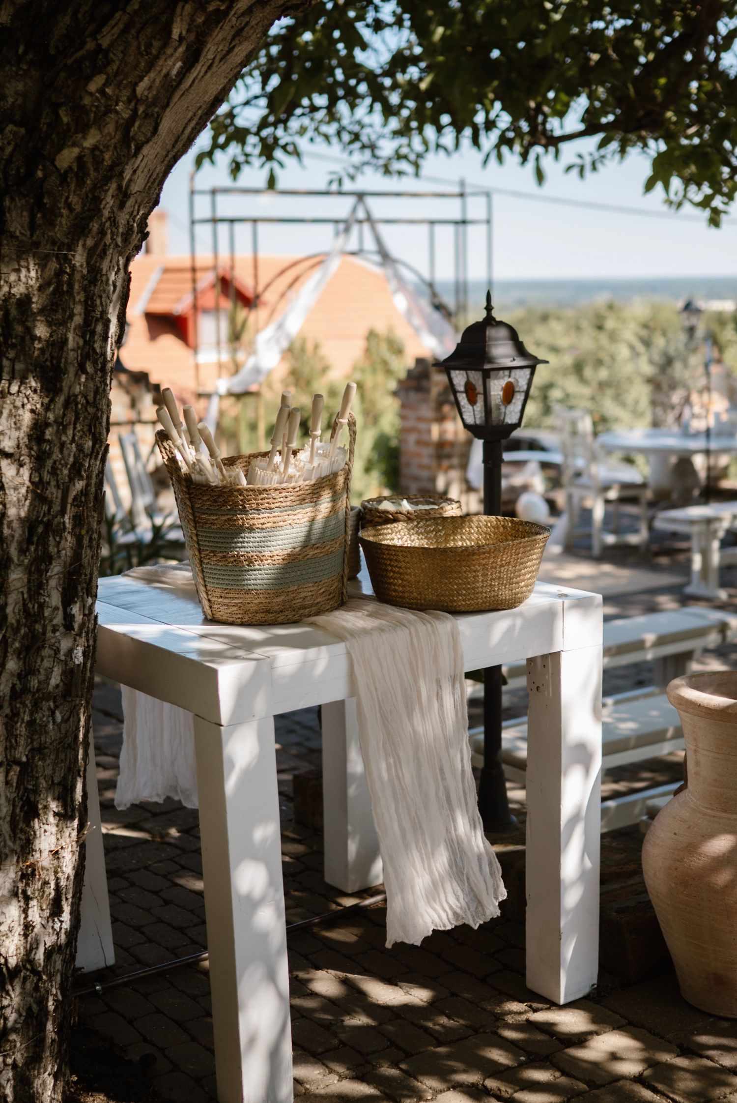 White table with woven baskets and lantern outdoors at a rustic wedding venue under a tree.