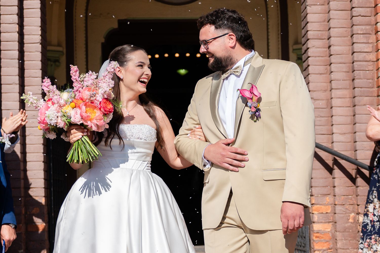 Joyful wedding party celebrates on steps of pink building with string lights and floral bouquets.